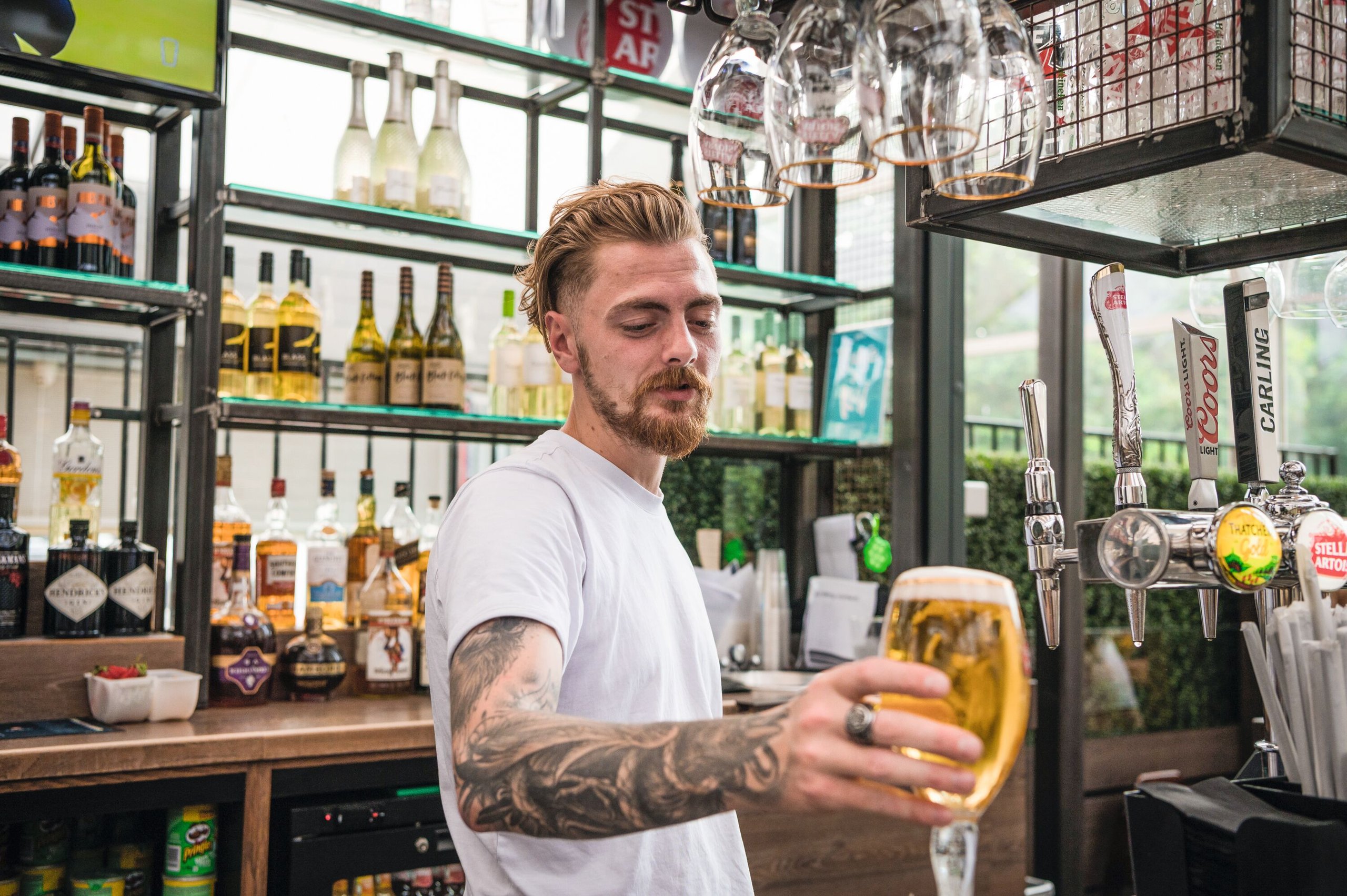 Bartender with tattoos on his arm holds up a pint of beer in front of shelves stocked with liquor bottles and beer taps.