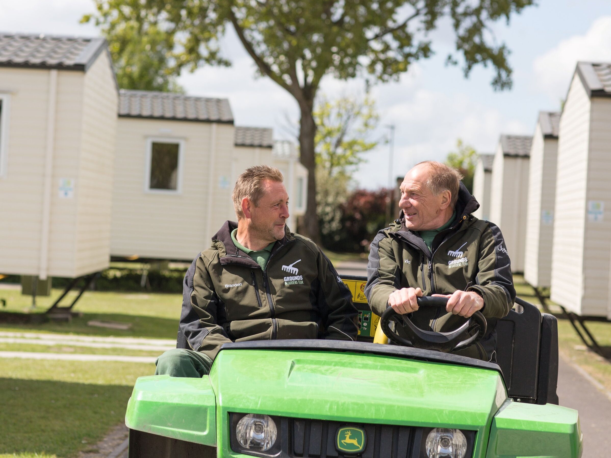 Two men in matching "Grounds Gardens Team" jackets sit in a green utility vehicle, smiling and talking to each other.