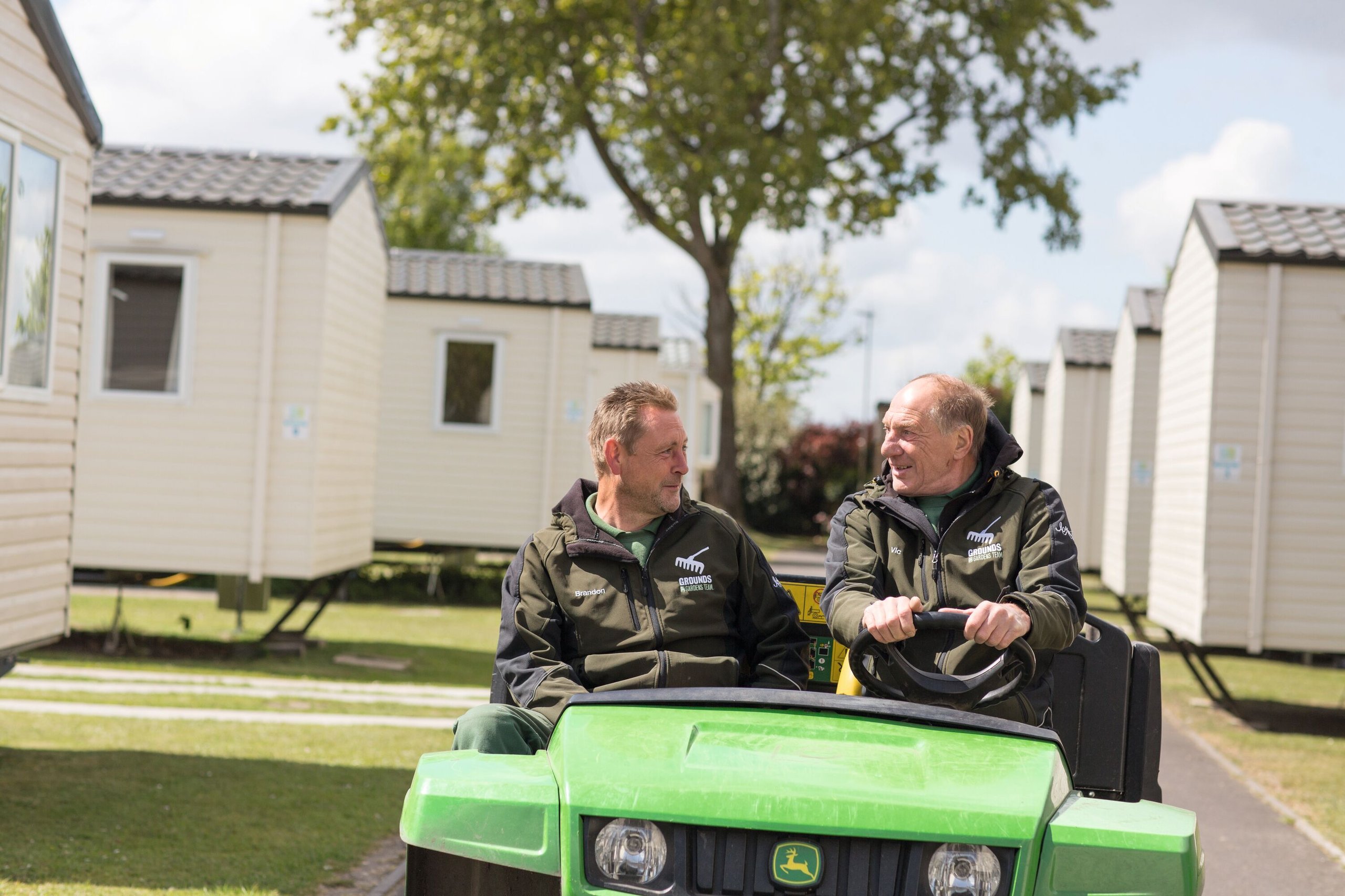 Two men in matching "Grounds Gardens Team" jackets sit in a green utility vehicle, smiling and talking to each other.