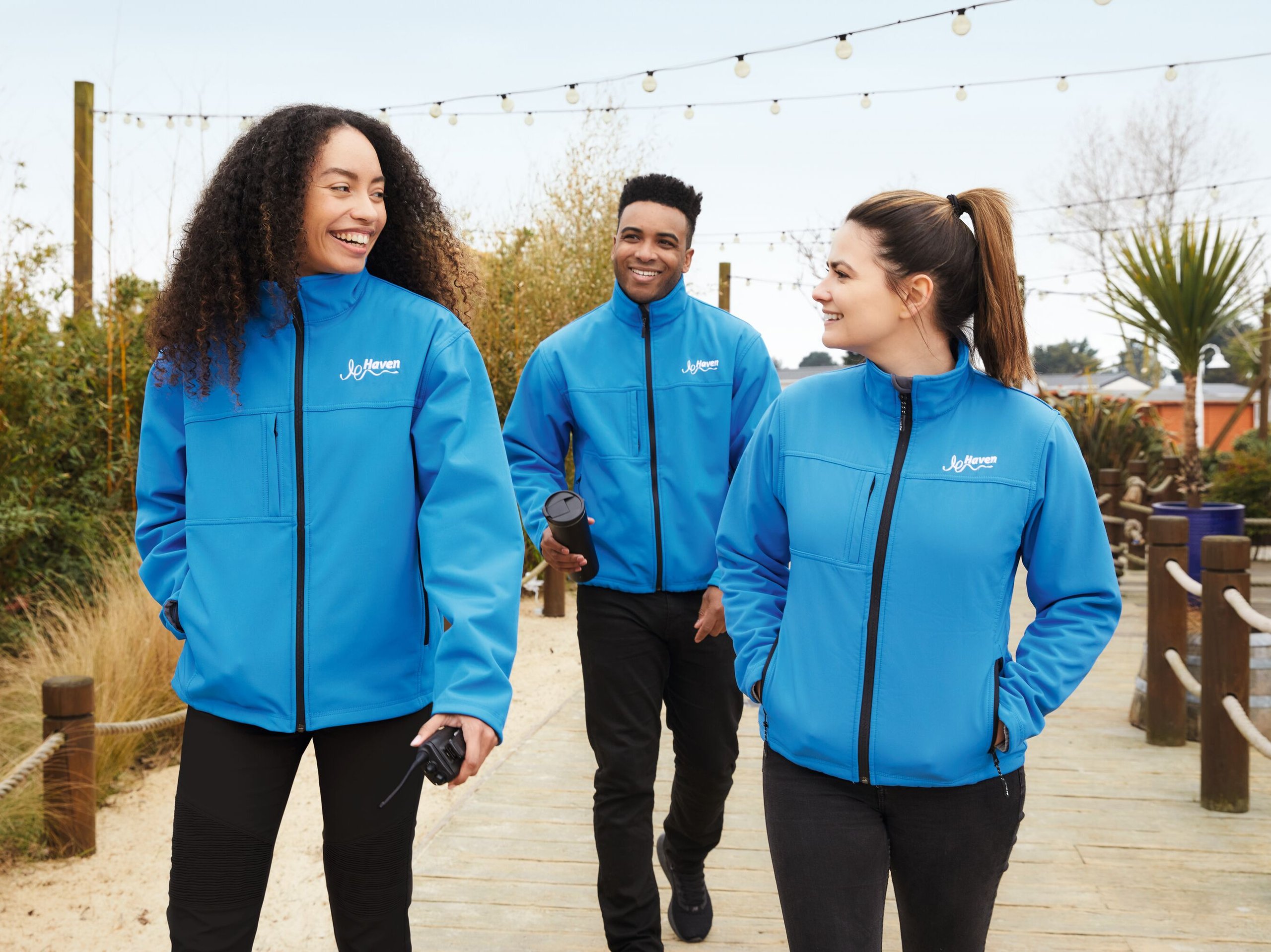 Three smiling people in blue Haven jackets walk on a wooden path, outdoors.