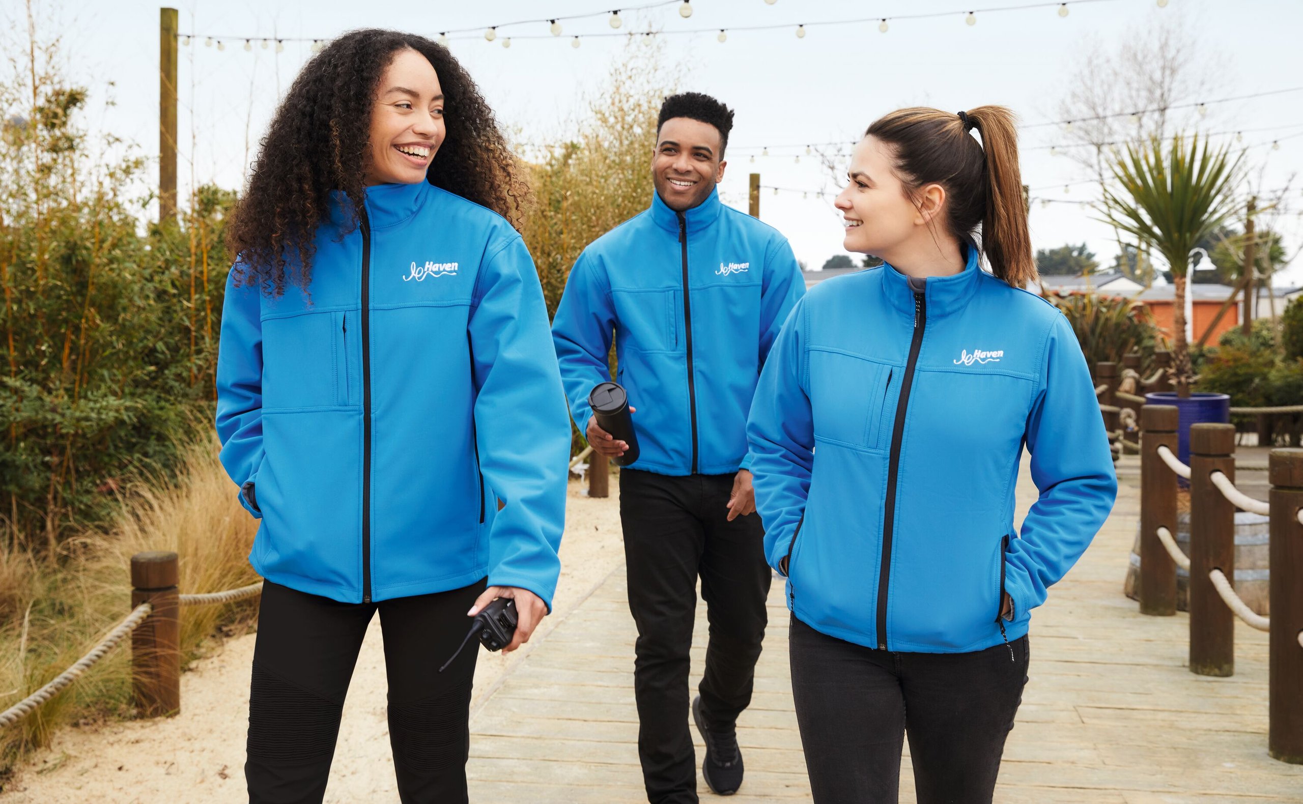 Three smiling people in blue Haven jackets walk on a wooden path, outdoors.