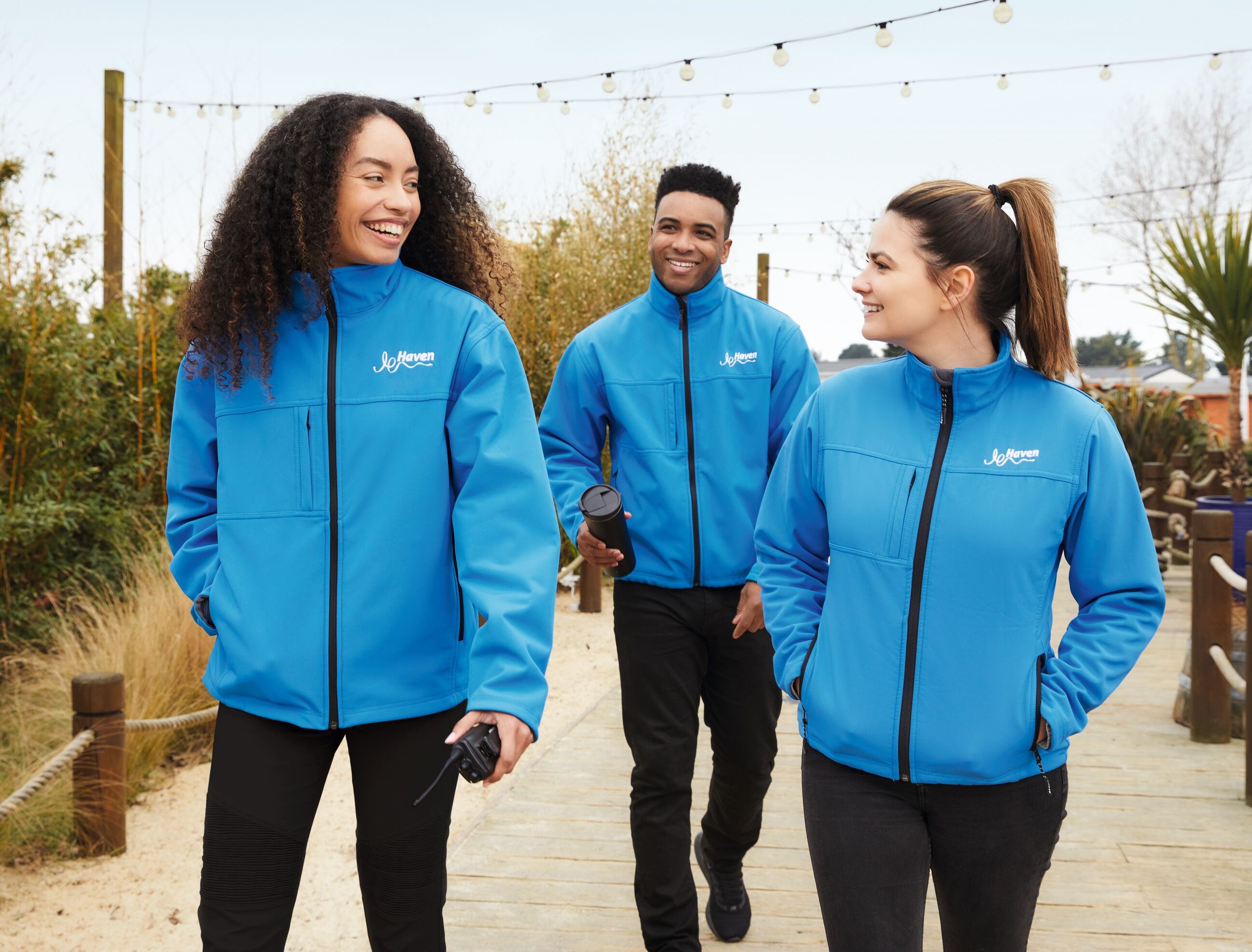 Three smiling people in blue Haven jackets walk on a wooden path, outdoors.