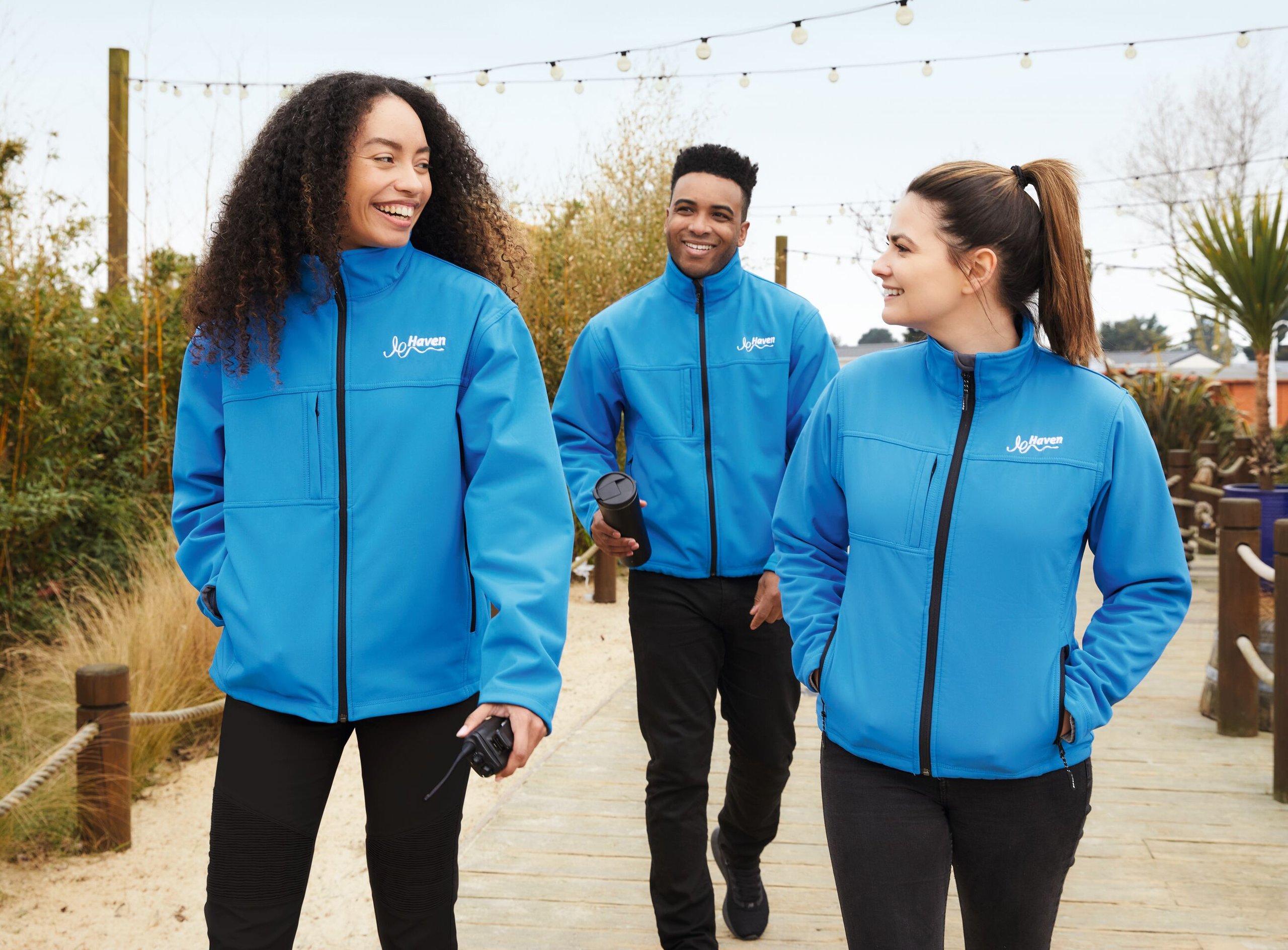 Three smiling people in blue Haven jackets walk on a wooden path, outdoors.