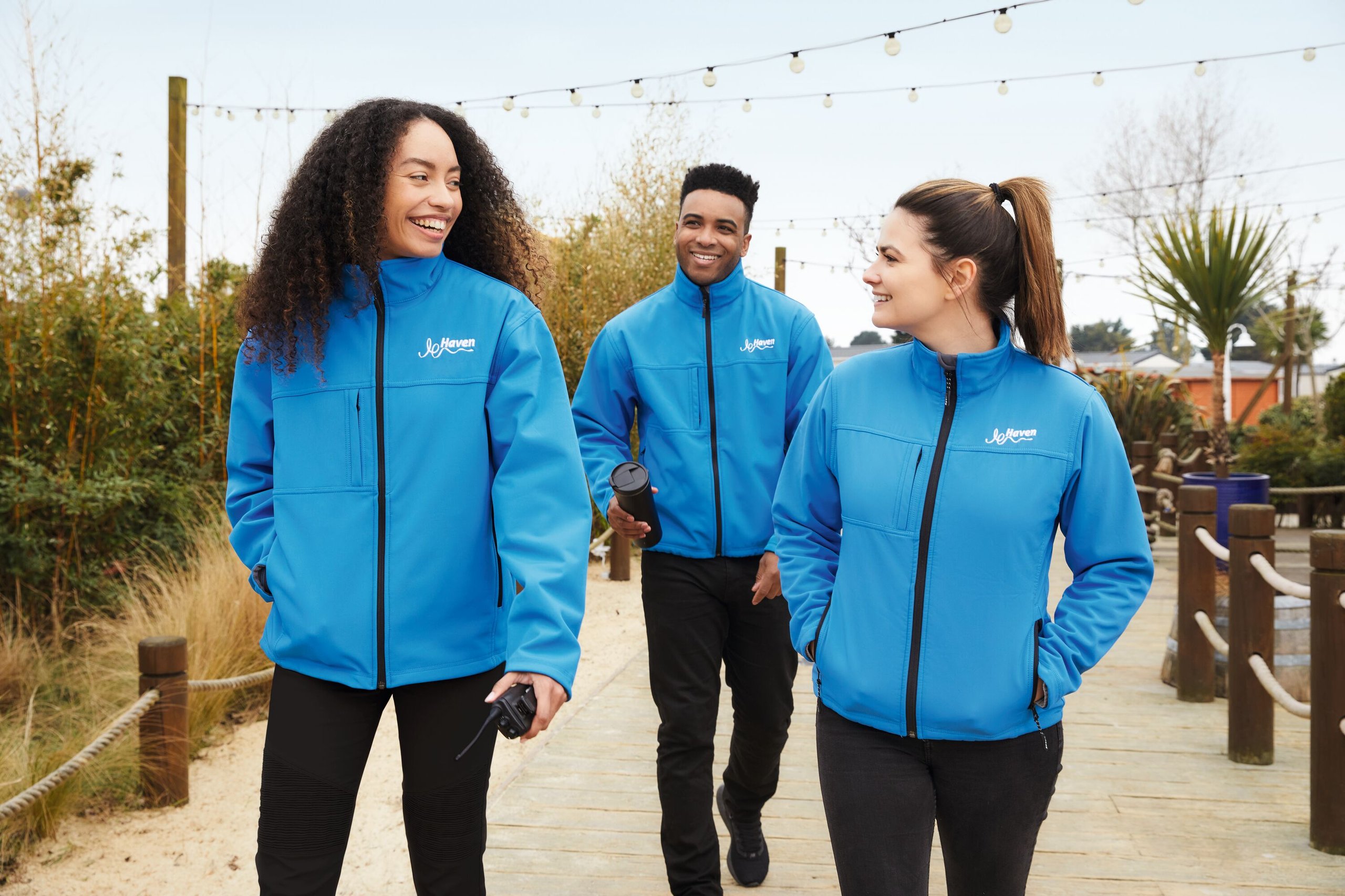 Three smiling people in blue Haven jackets walk on a wooden path, outdoors.