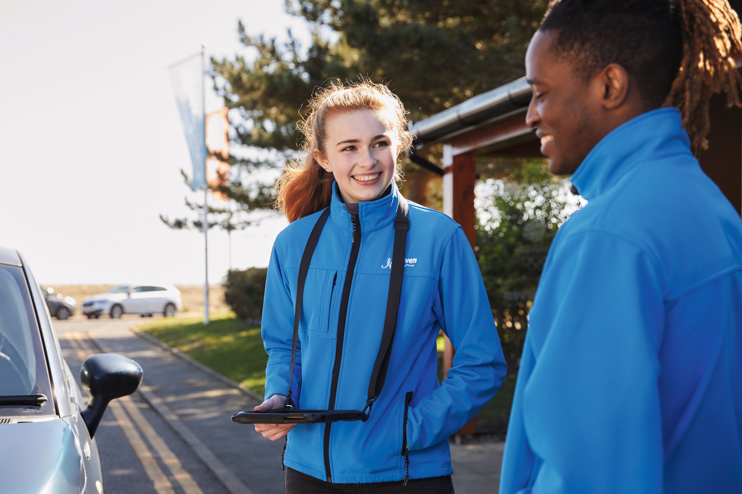 Two smiling people in blue jackets are outside with a car nearby. One holds a tablet and looks at the other.