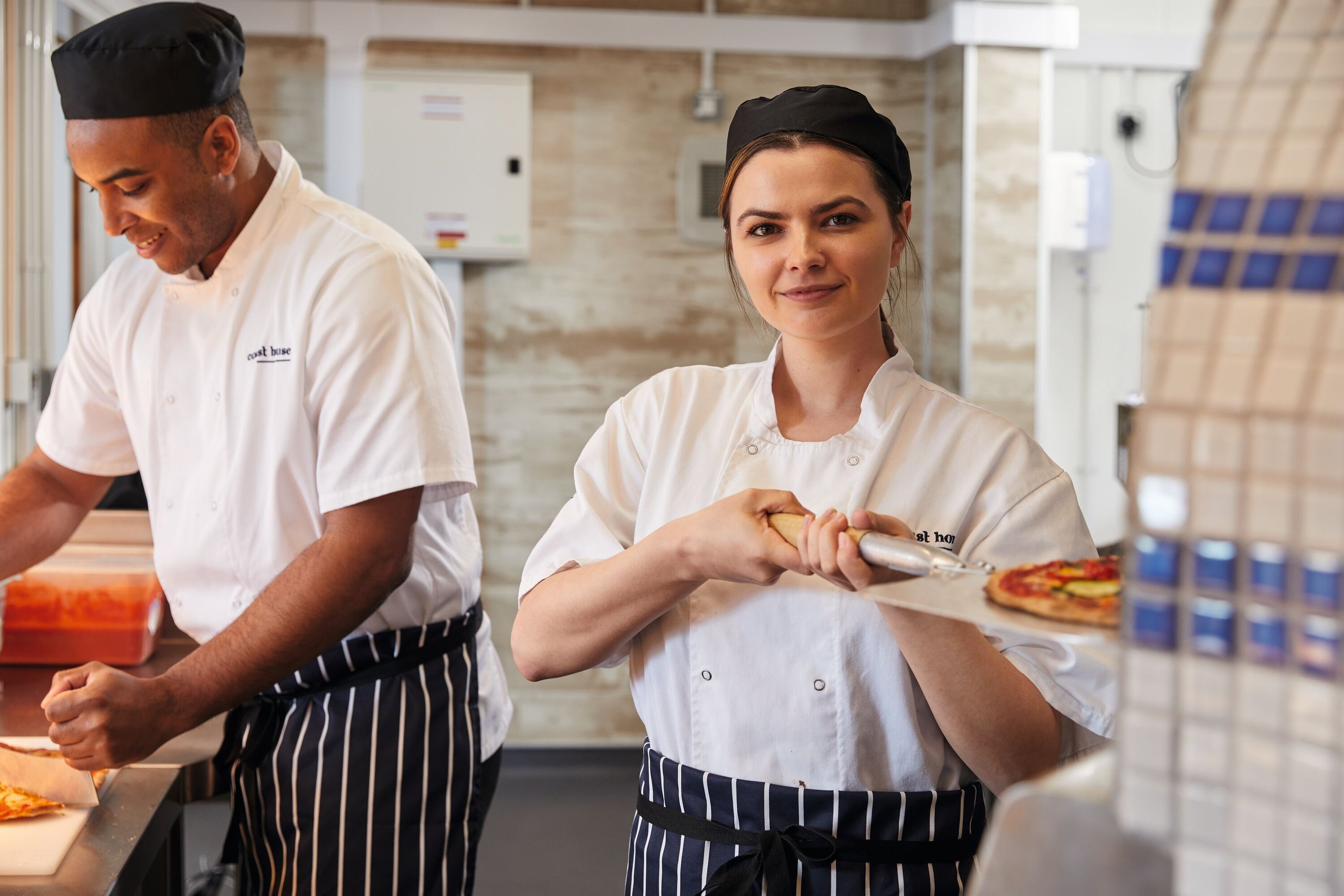 Two chefs, a man and a woman, wearing chef whites and black aprons, work in a kitchen. The woman holds a pizza peel with a pizza.