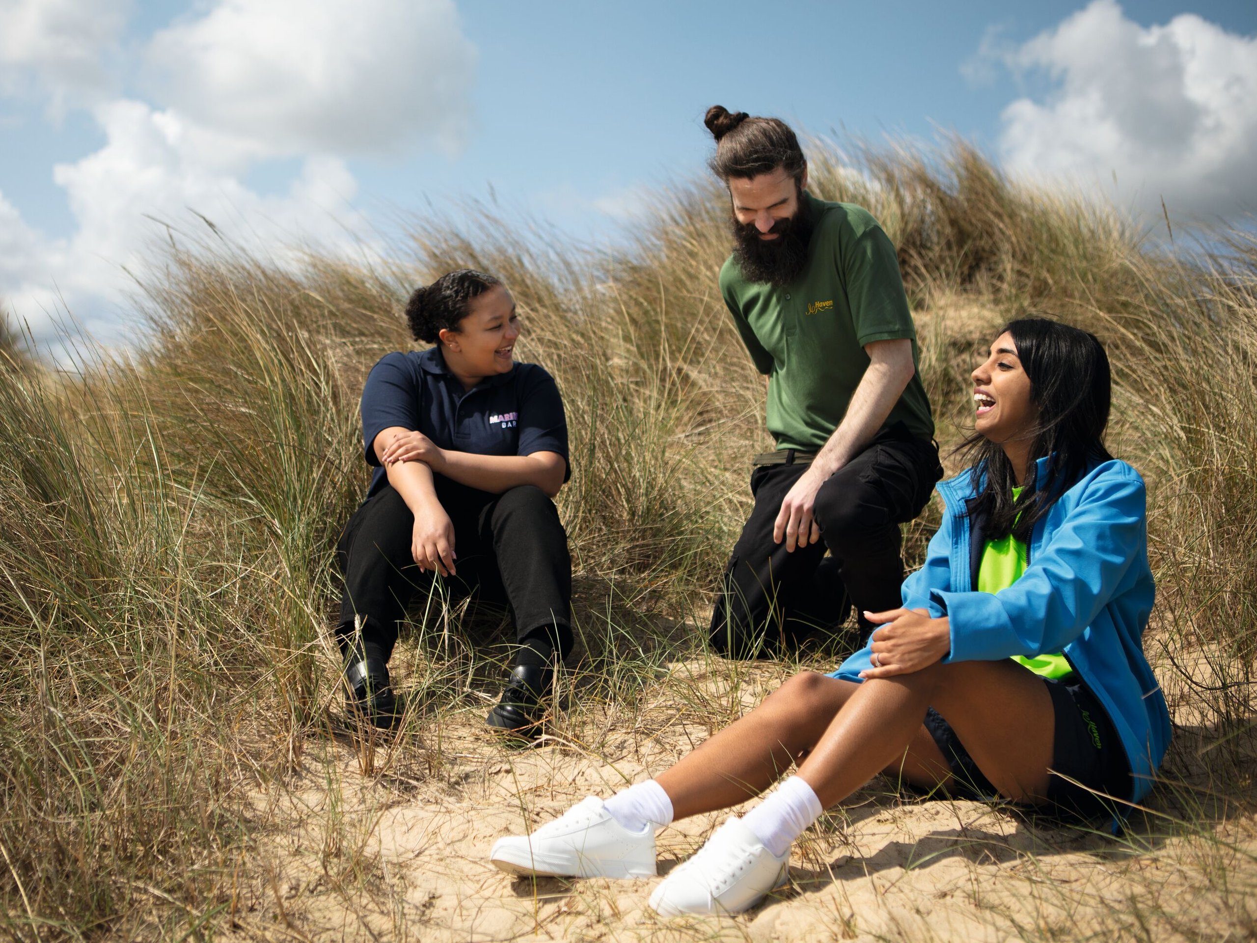 Three people, two women and one man, sit and chat amongst tall dune grasses under a blue sky with scattered clouds.