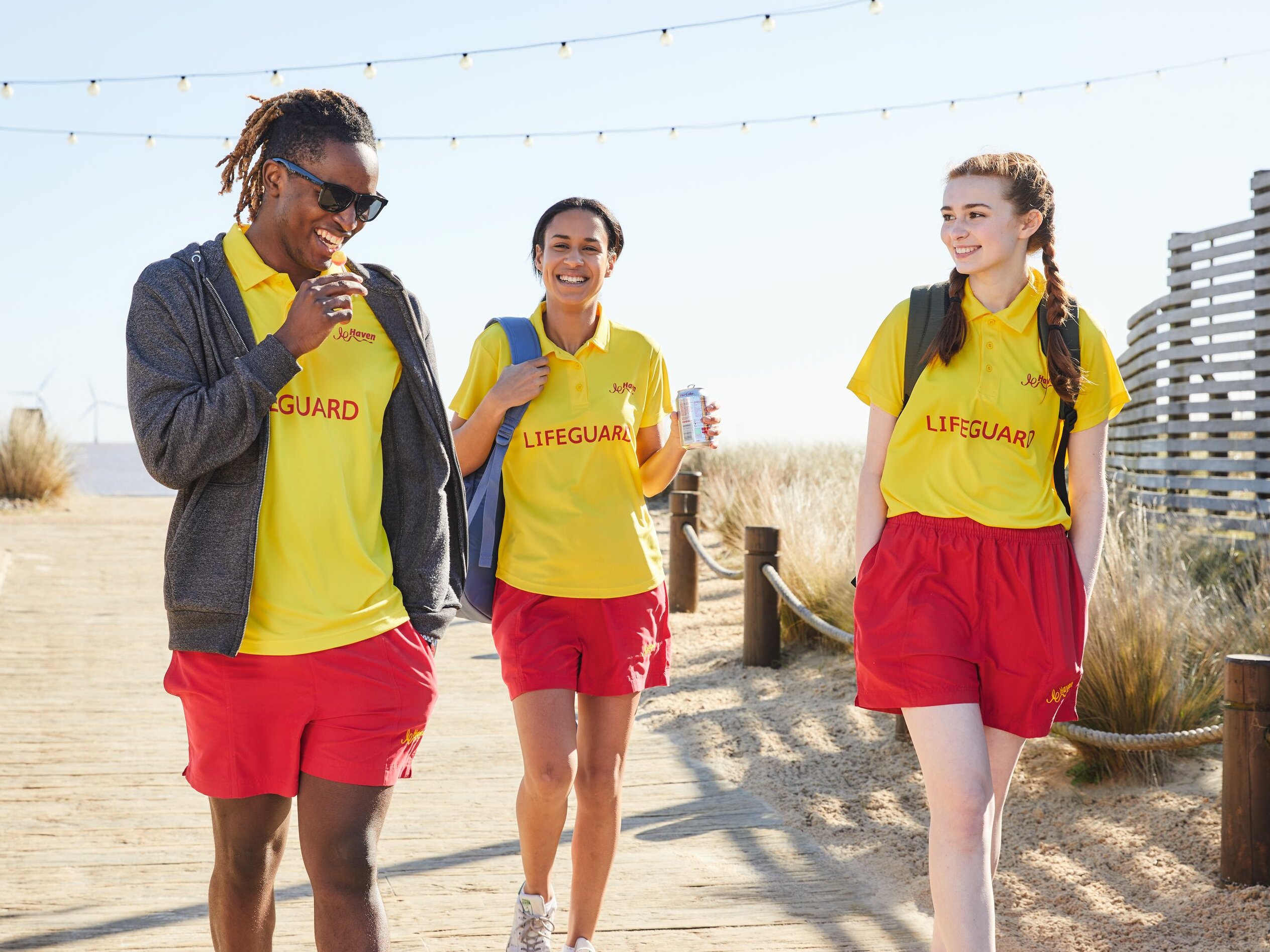 Three lifeguards in yellow shirts and red shorts walk on a wooden boardwalk, smiling.