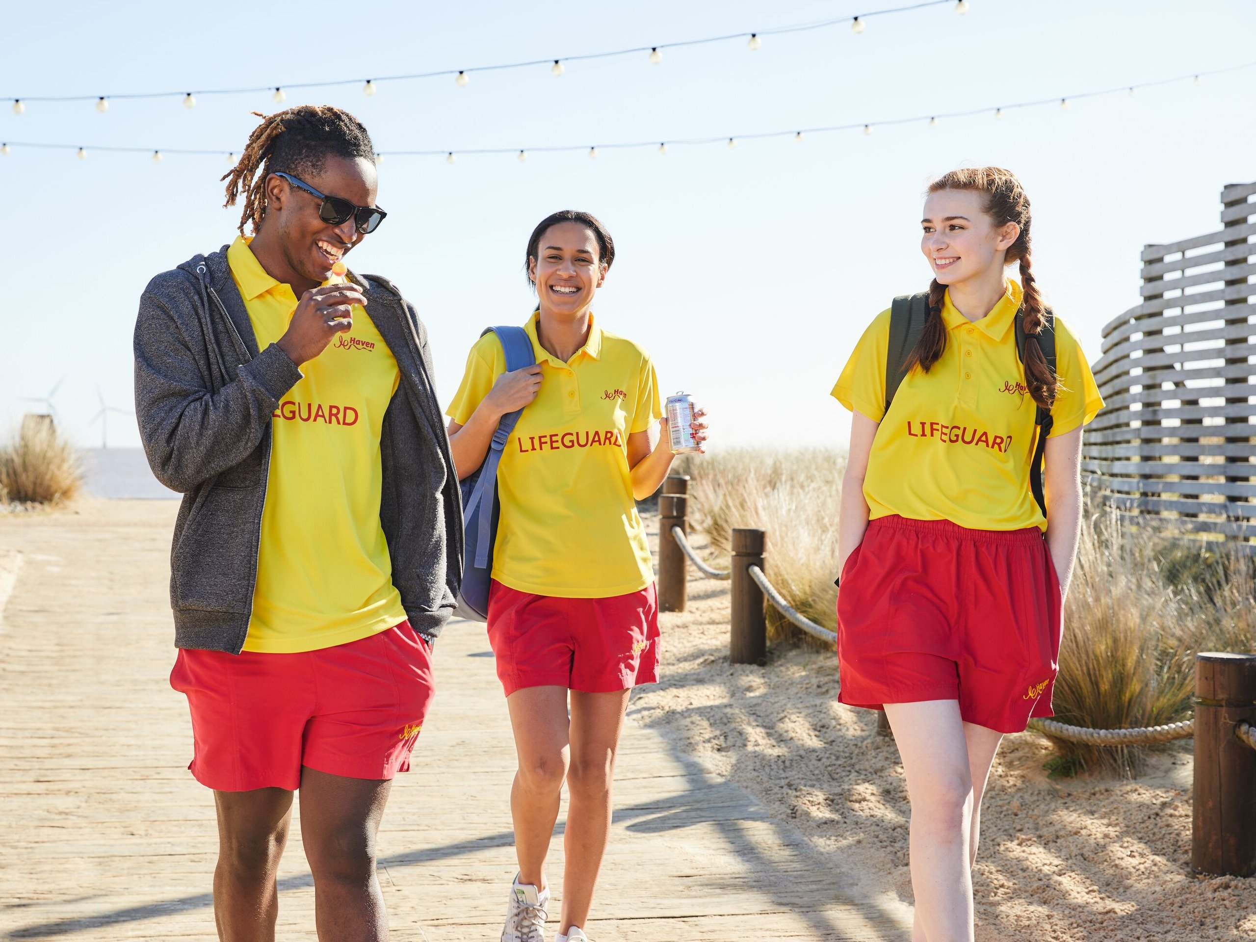 Three lifeguards in yellow shirts and red shorts walk on a wooden boardwalk, smiling.