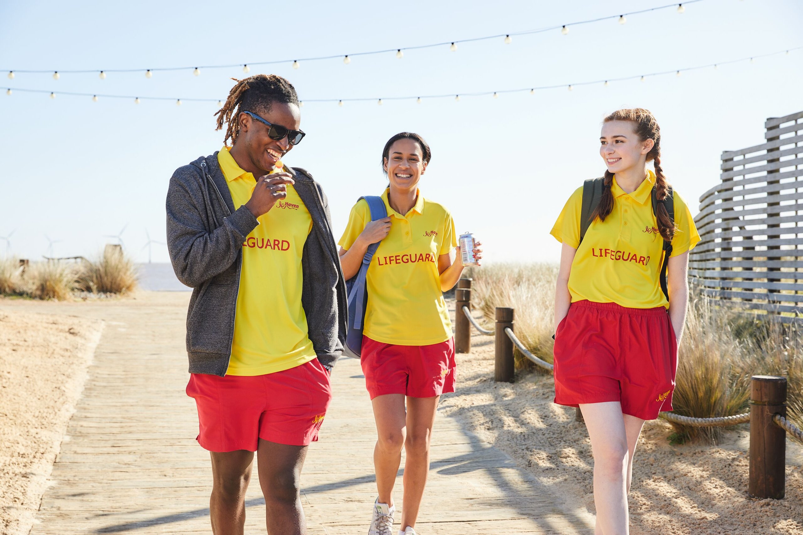 Three lifeguards in yellow shirts and red shorts walk on a wooden boardwalk, smiling.