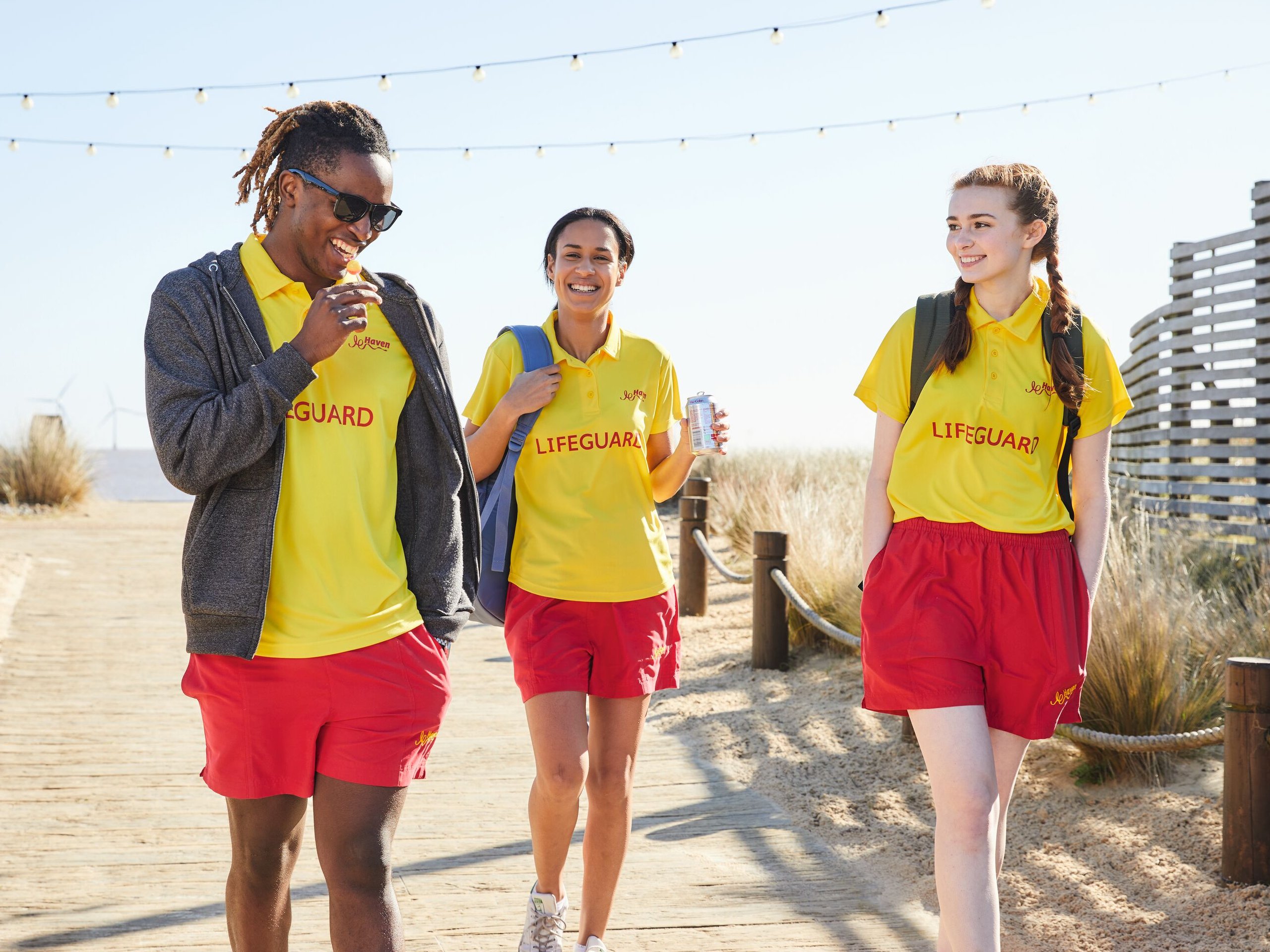Three lifeguards in yellow shirts and red shorts walk on a wooden boardwalk, smiling.