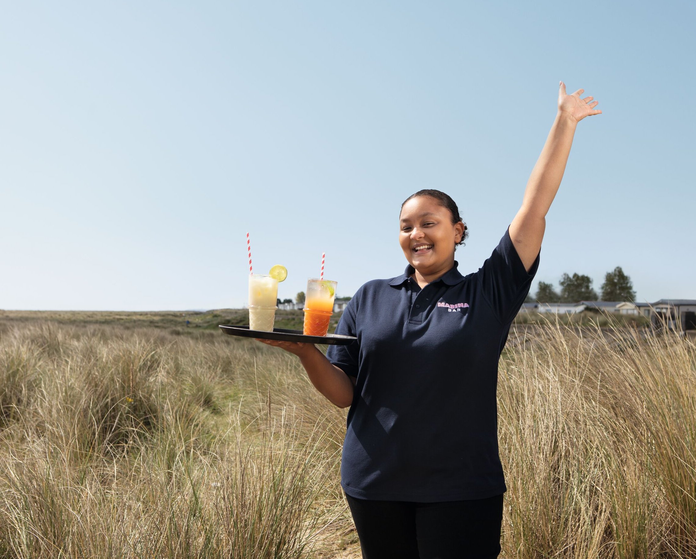 A smiling woman in a navy blue polo shirt with "Marina Bar" on it holds a tray with two cocktails. She stands in tall grass with her arm raised.