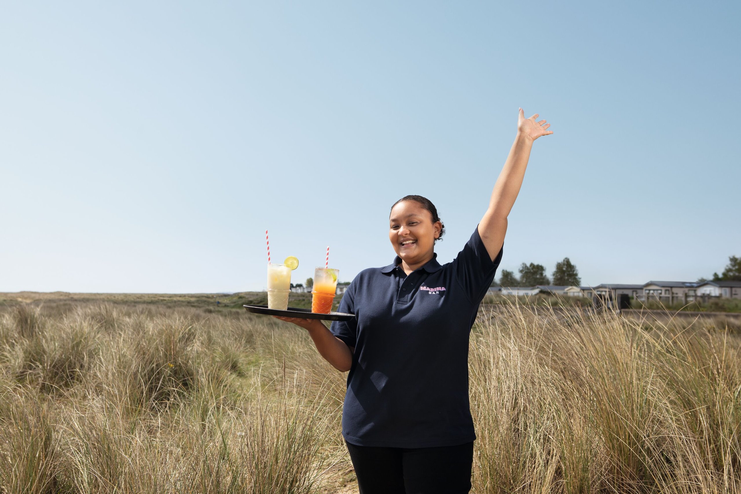 A smiling woman in a navy blue polo shirt with "Marina Bar" on it holds a tray with two cocktails. She stands in tall grass with her arm raised.