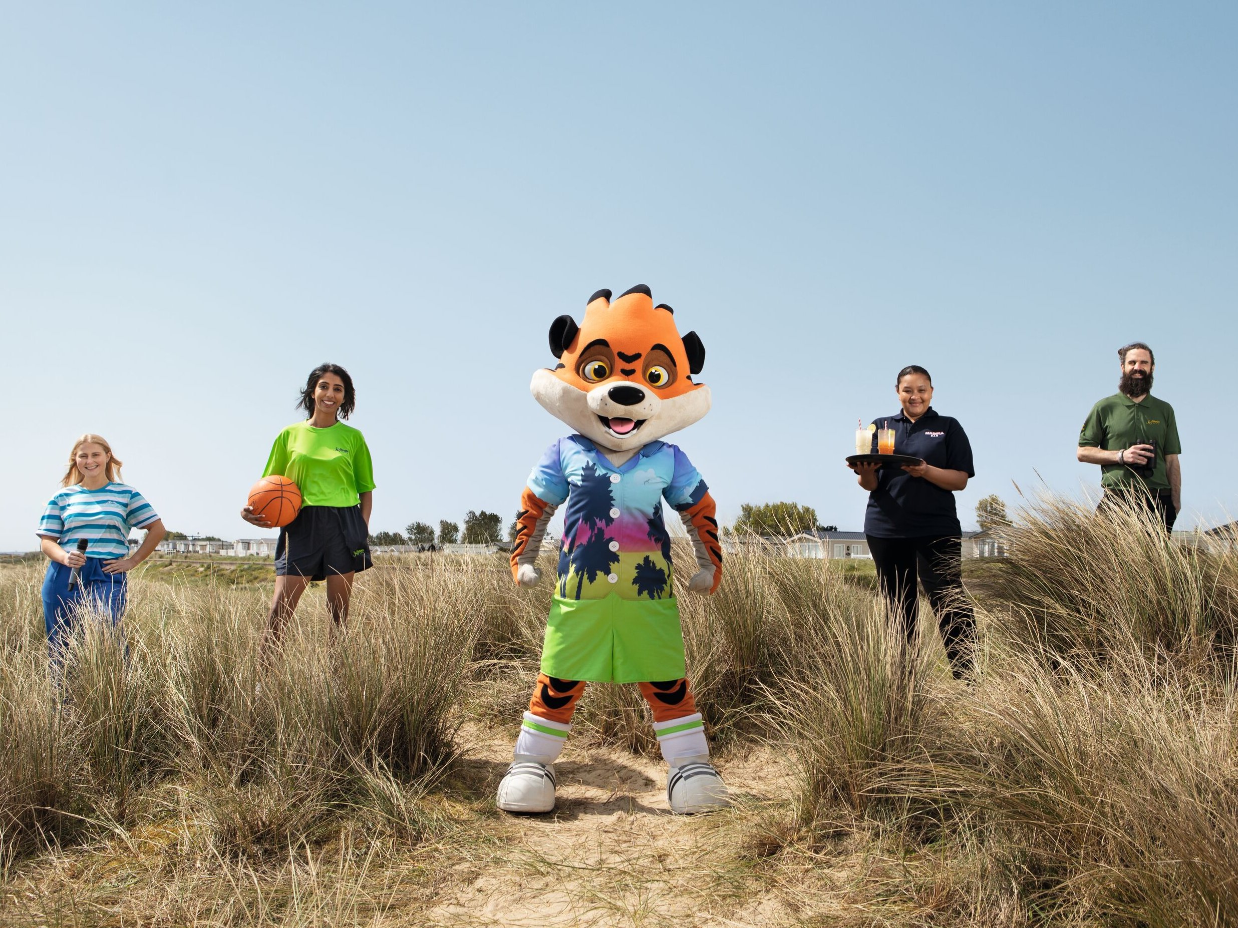 A mascot and four staff members stand in tall grass with a clear blue sky. One staff member holds a basketball, another offers drinks, and a third holds binoculars.