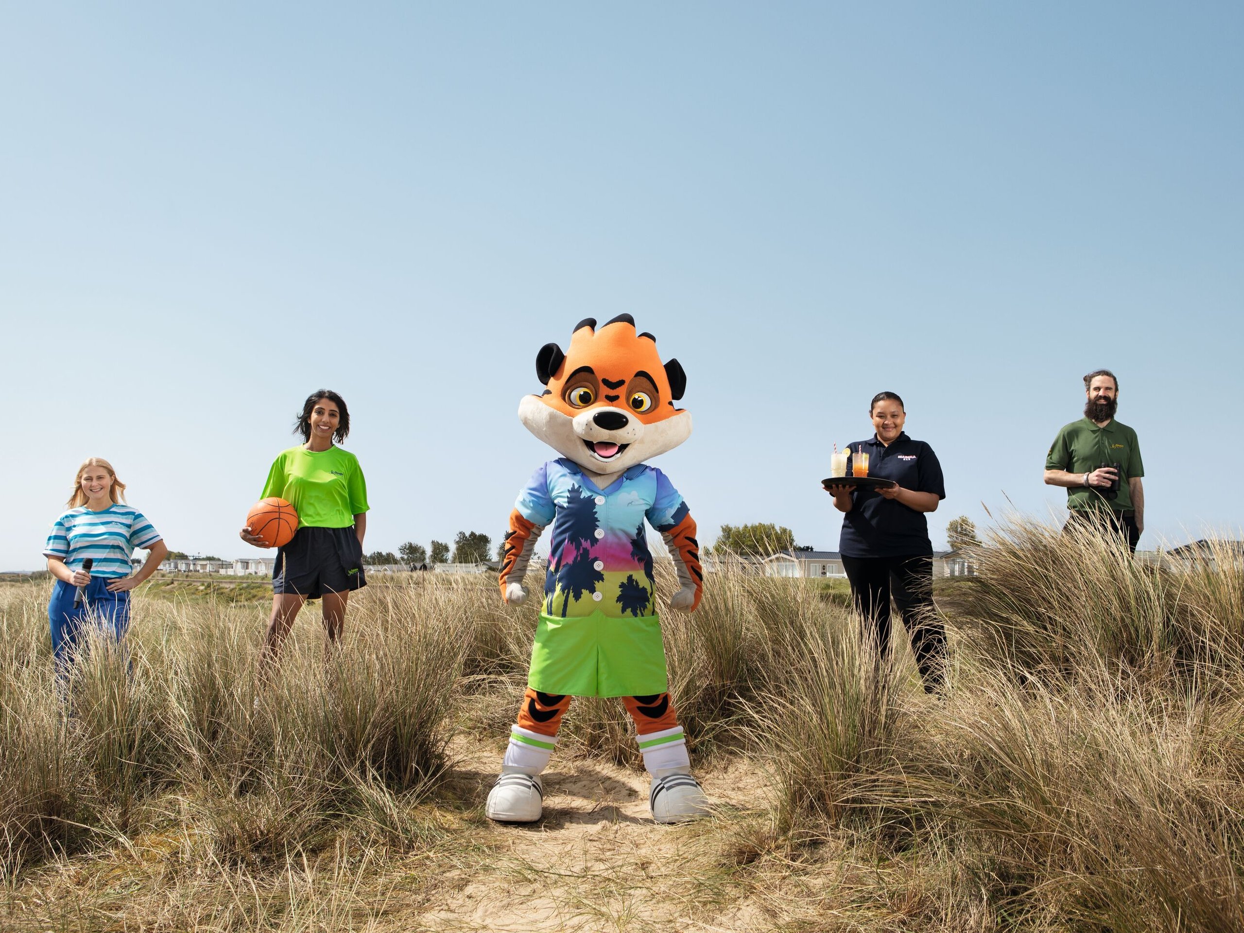 A mascot and four staff members stand in tall grass with a clear blue sky. One staff member holds a basketball, another offers drinks, and a third holds binoculars.