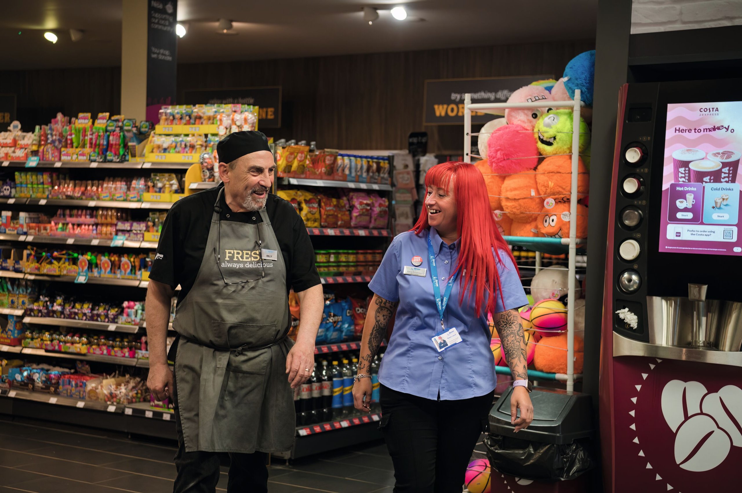 A male store worker in an apron and a female worker with bright red hair chat while standing in an aisle of a convenience store.