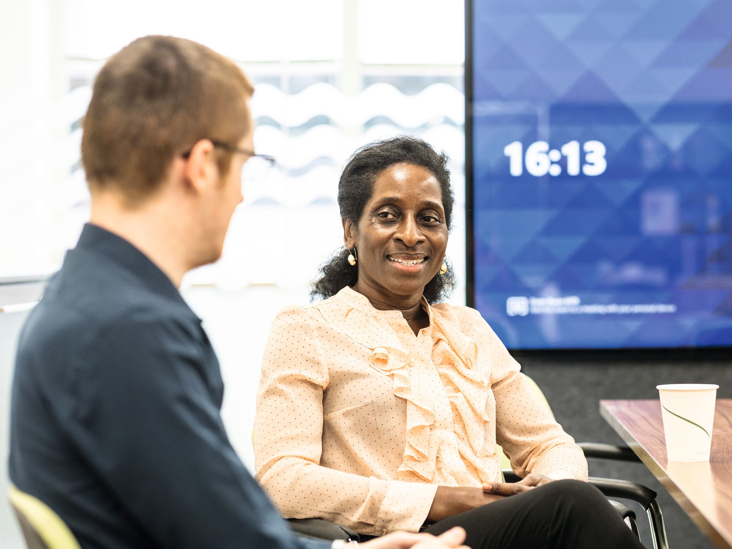 Two colleagues, a Black woman and a white man, conversing in a meeting room with a digital display showing the time 16:13.
