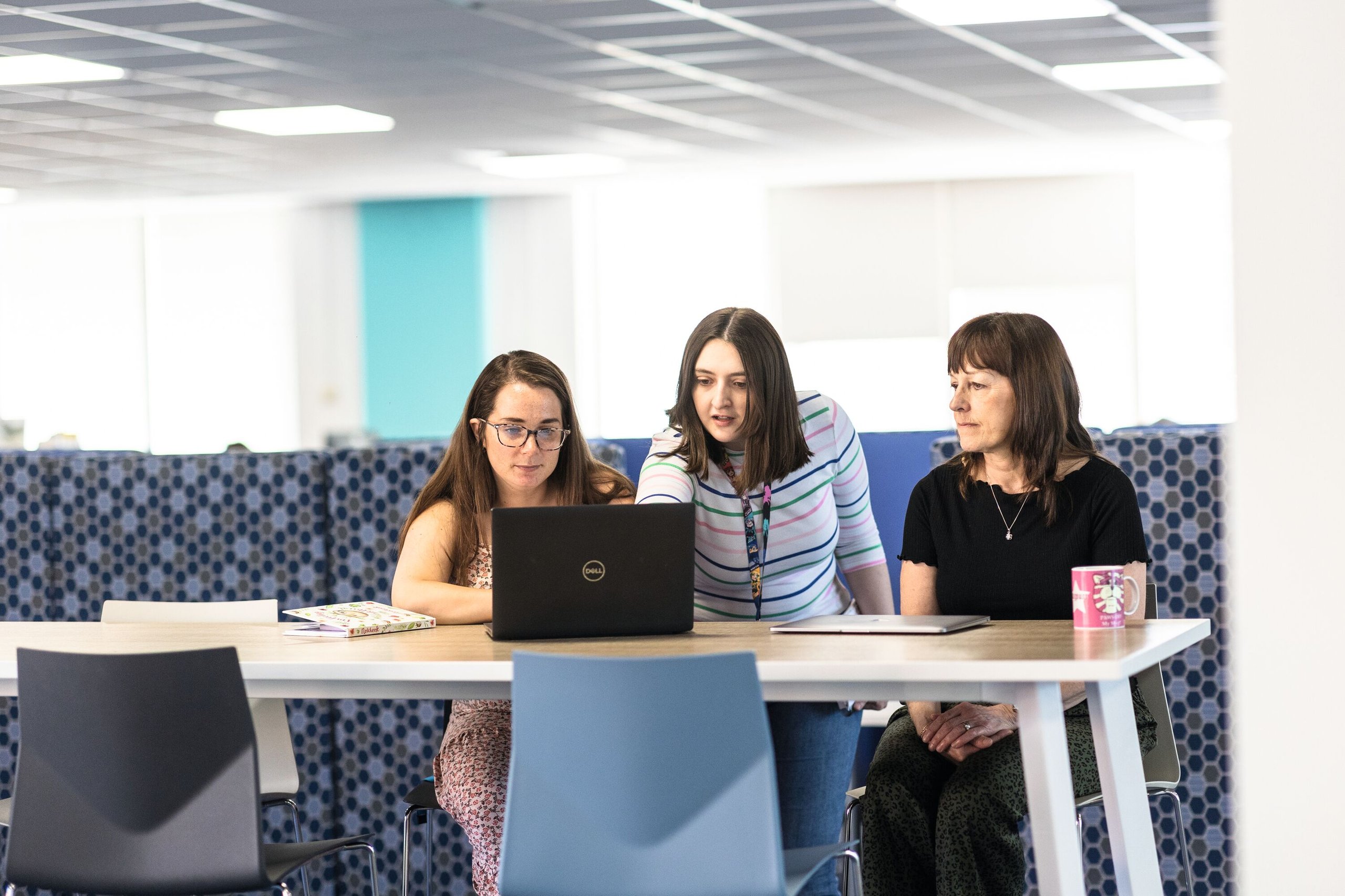 Three women huddle around a laptop at a table in a modern office, with one pointing at the screen.