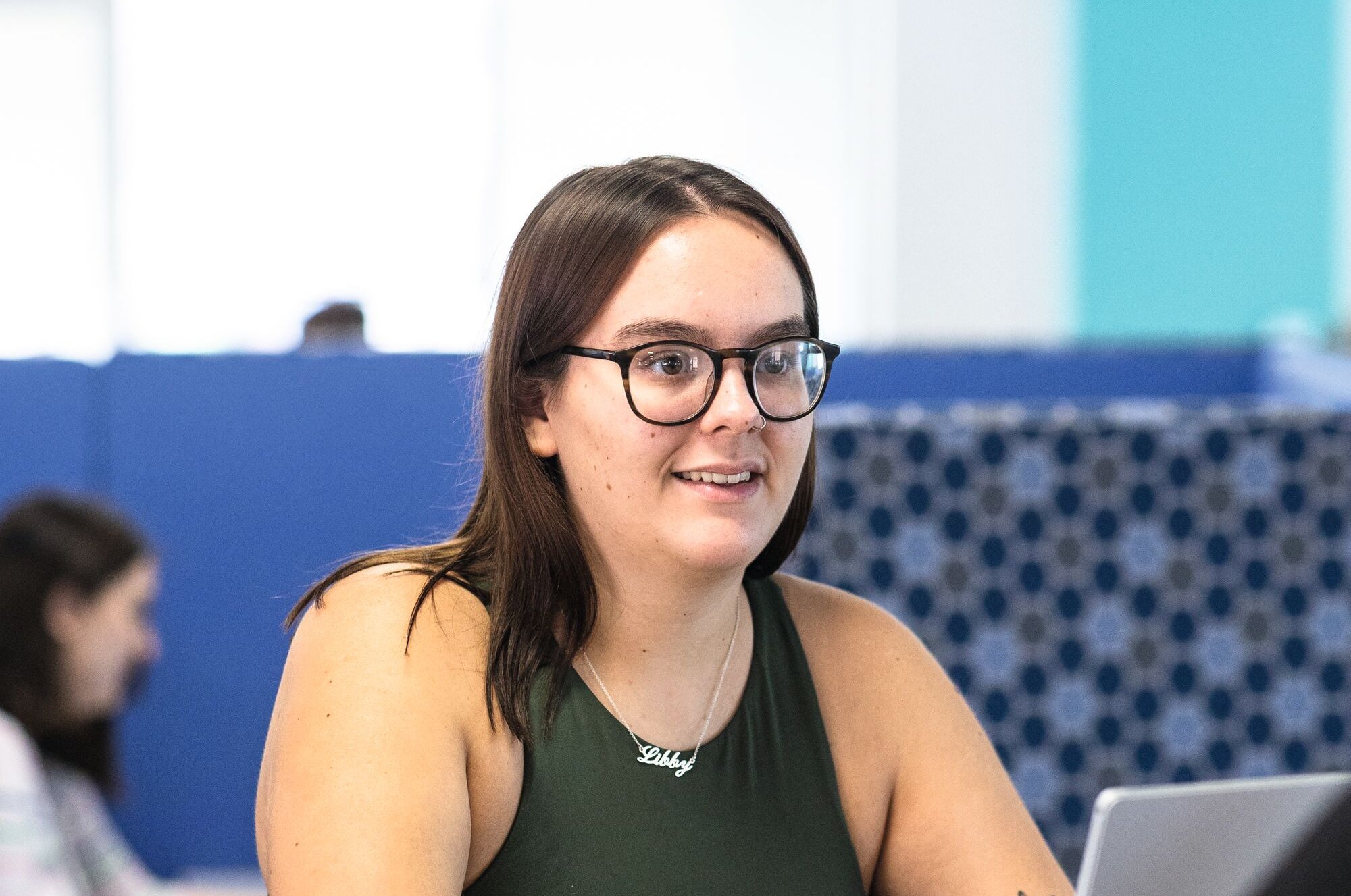 A young woman wearing glasses and a green top sits at a table, looking to the side. She wears a necklace with her name, "Libby," and has a butterfly tattoo on her arm.
