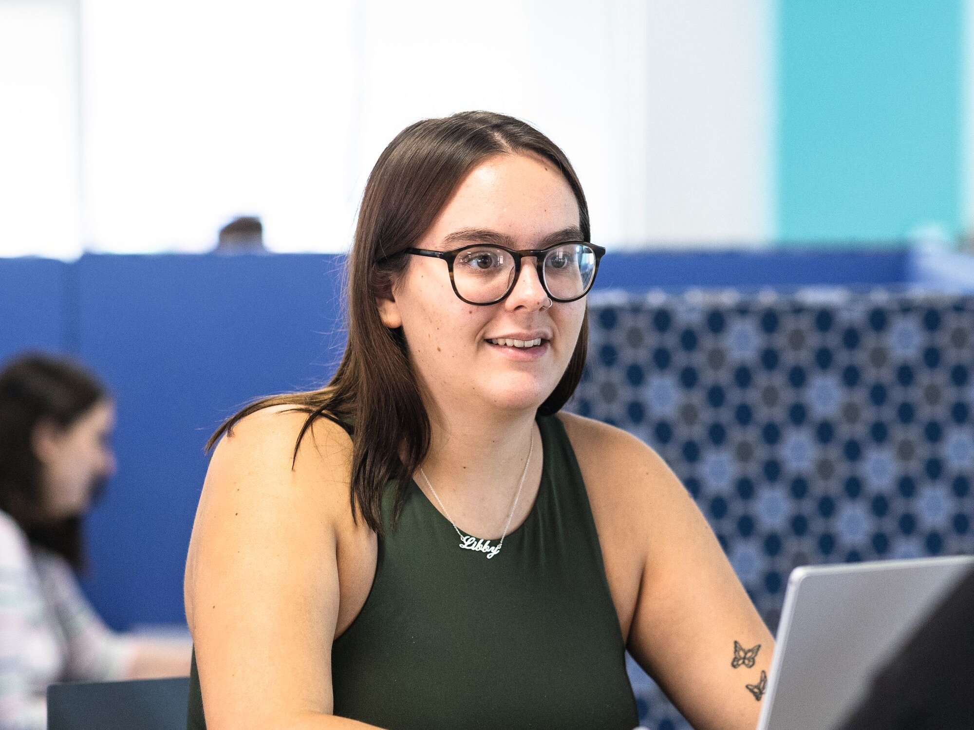 A young woman wearing glasses and a green top sits at a table, looking to the side. She wears a necklace with her name, "Libby," and has a butterfly tattoo on her arm.