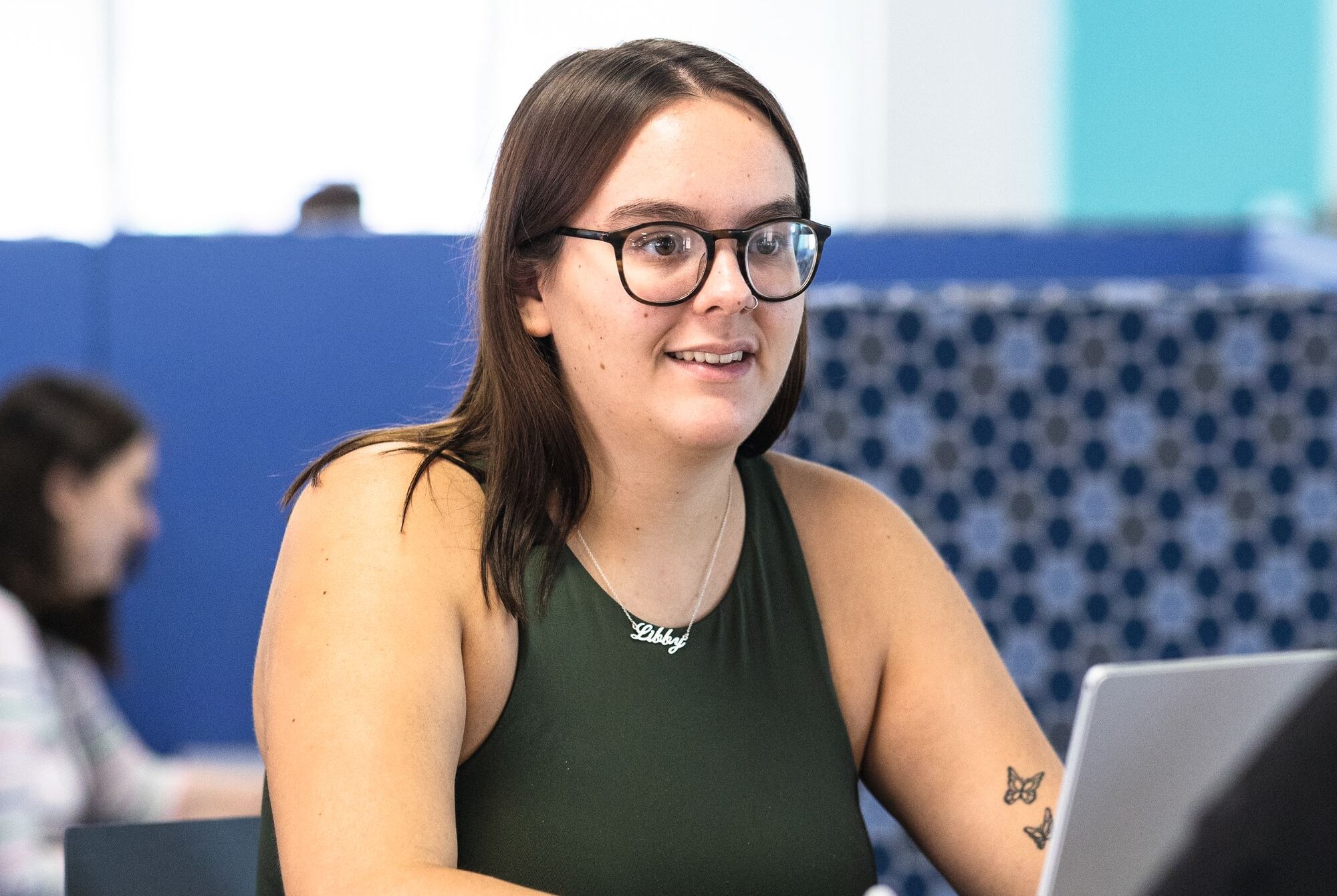 A young woman wearing glasses and a green top sits at a table, looking to the side. She wears a necklace with her name, "Libby," and has a butterfly tattoo on her arm.