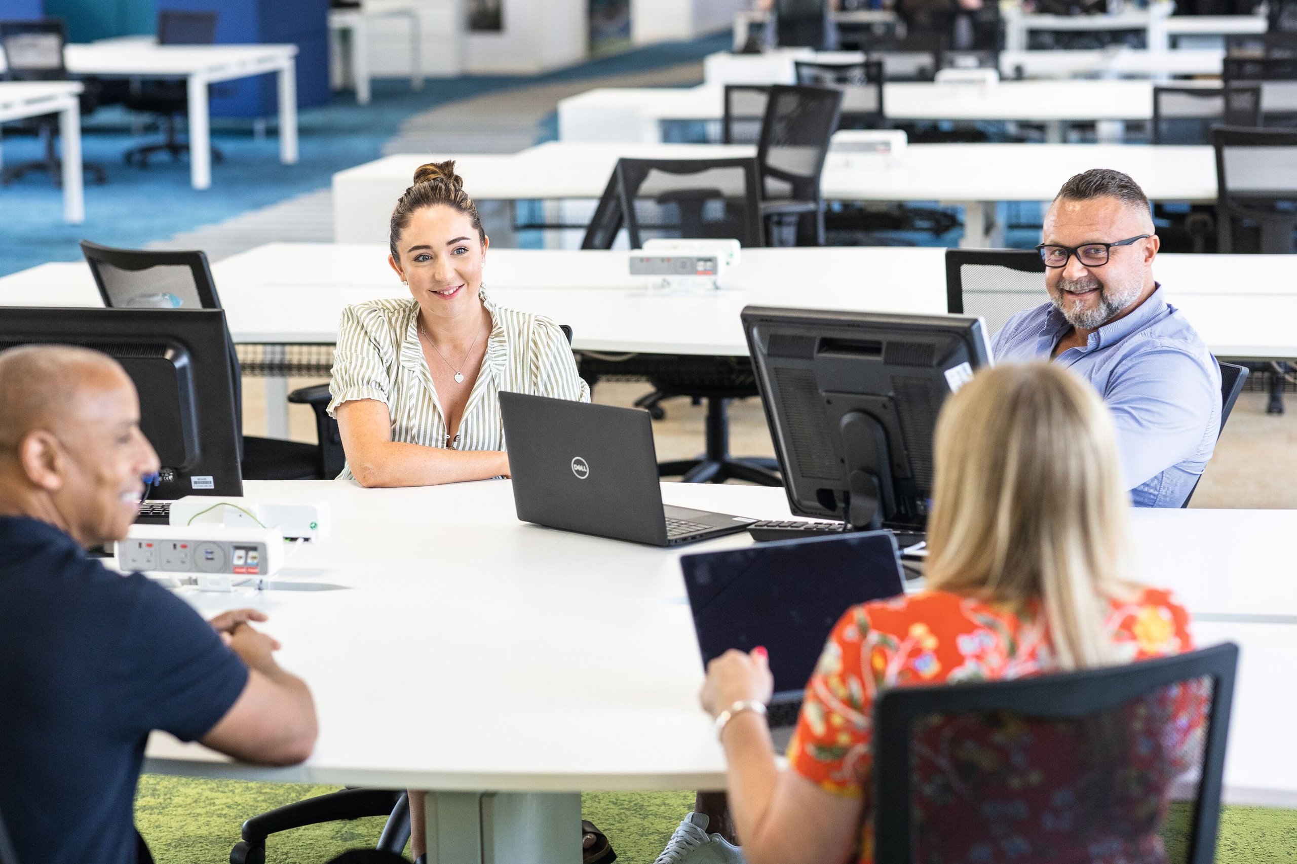 Four people are gathered around a white table in a modern office, working together on laptops and computer monitors.