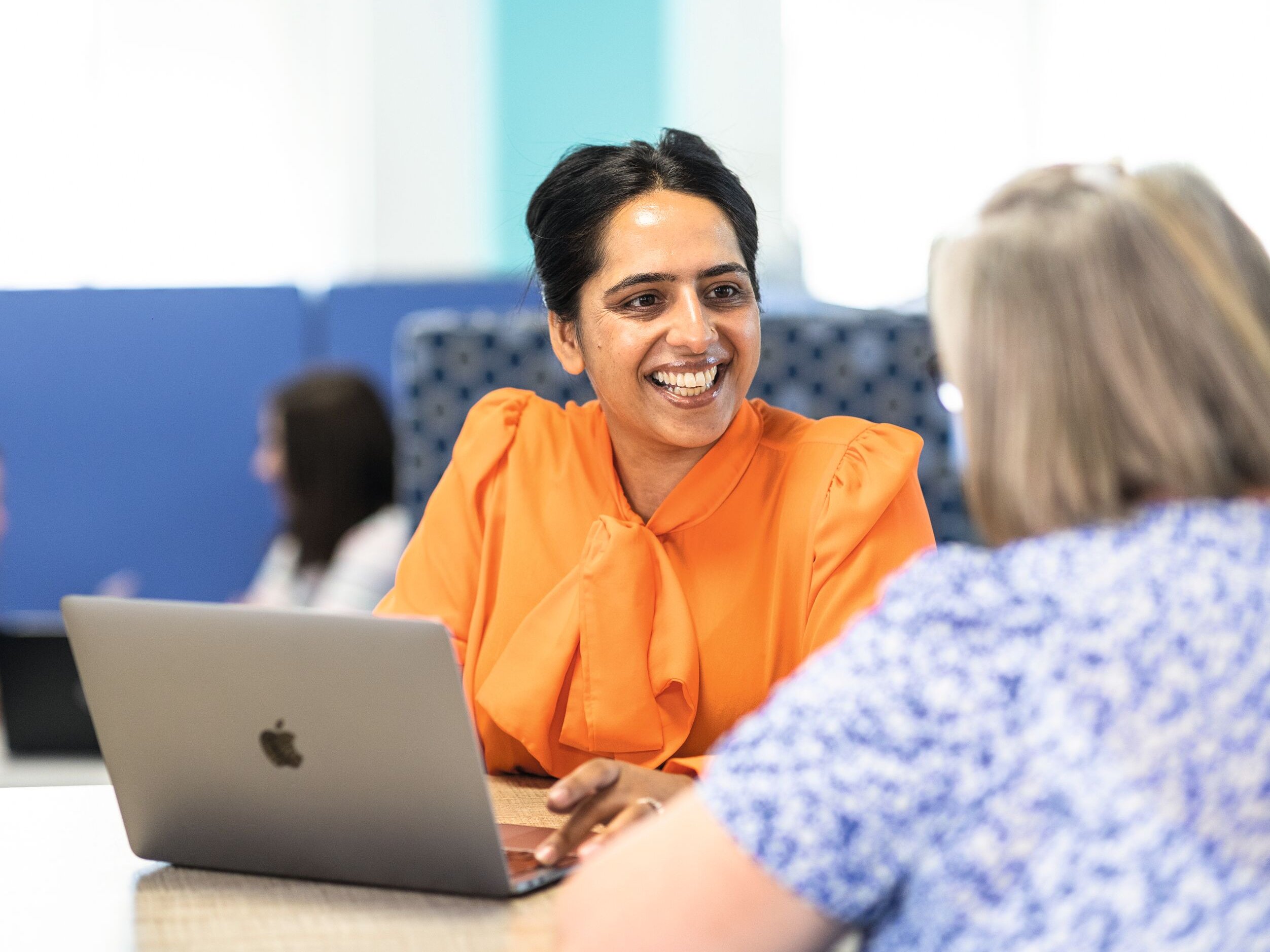 Two women are working together at a desk with a laptop. One woman is wearing an orange shirt and smiling.