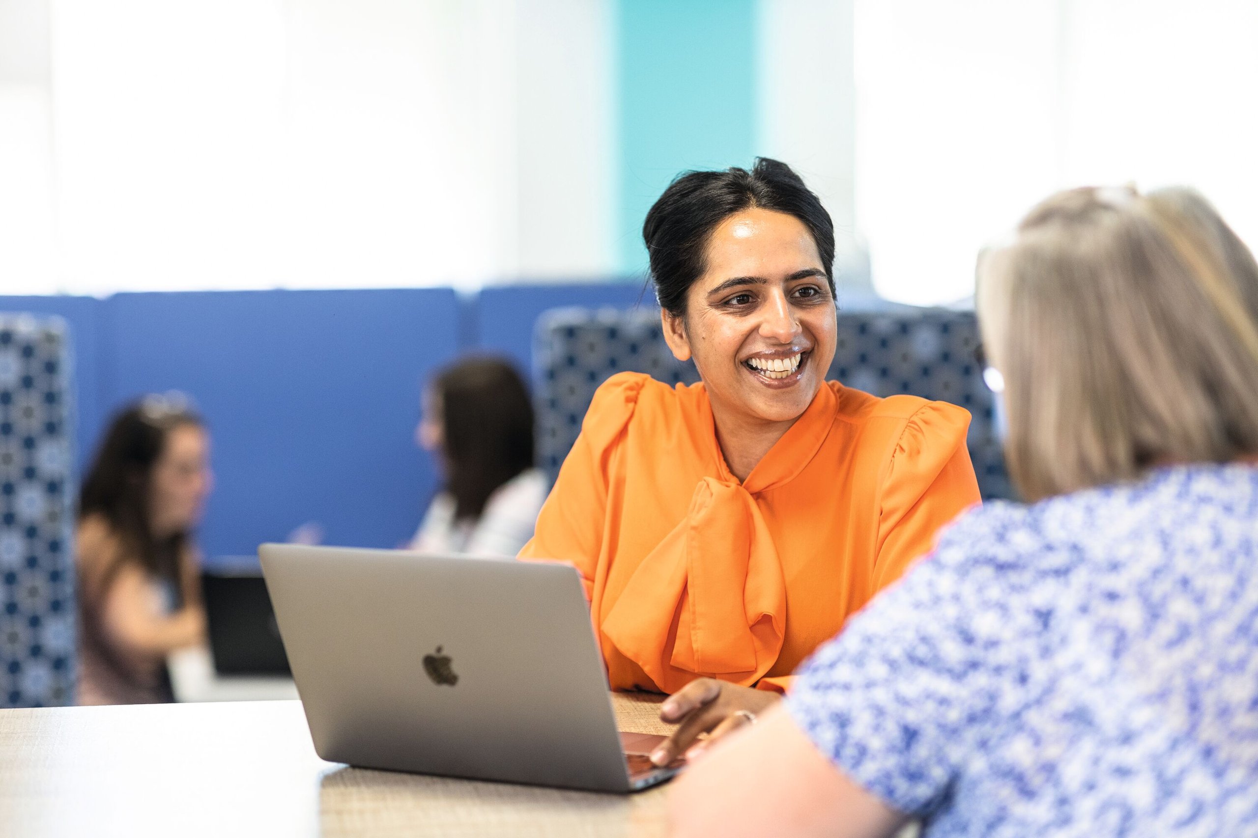 Two women are working together at a desk with a laptop. One woman is wearing an orange shirt and smiling.