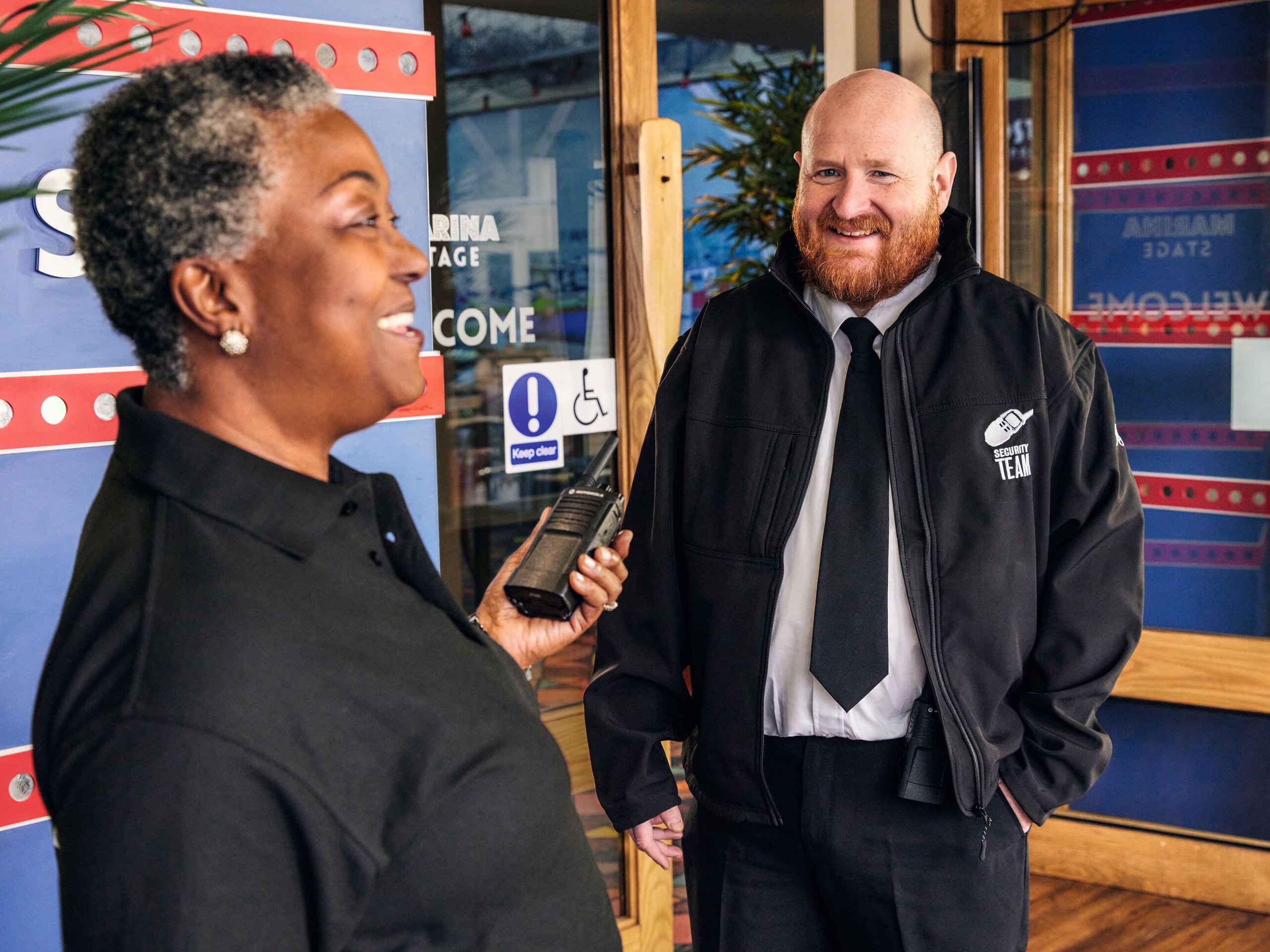 A female security guard holding a radio speaks with a male security guard in a jacket with "Security Team" on it.