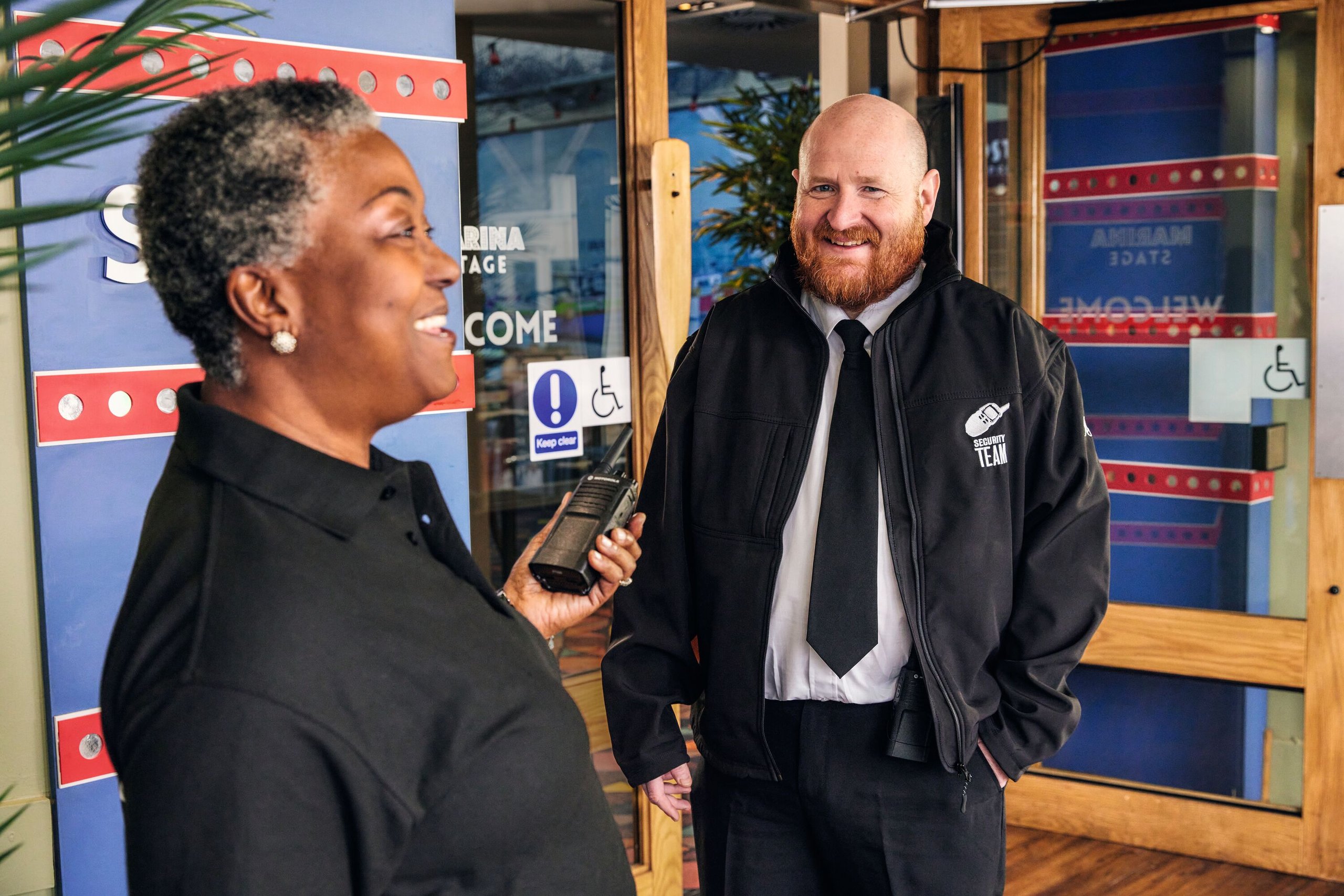 A female security guard holding a radio speaks with a male security guard in a jacket with "Security Team" on it.