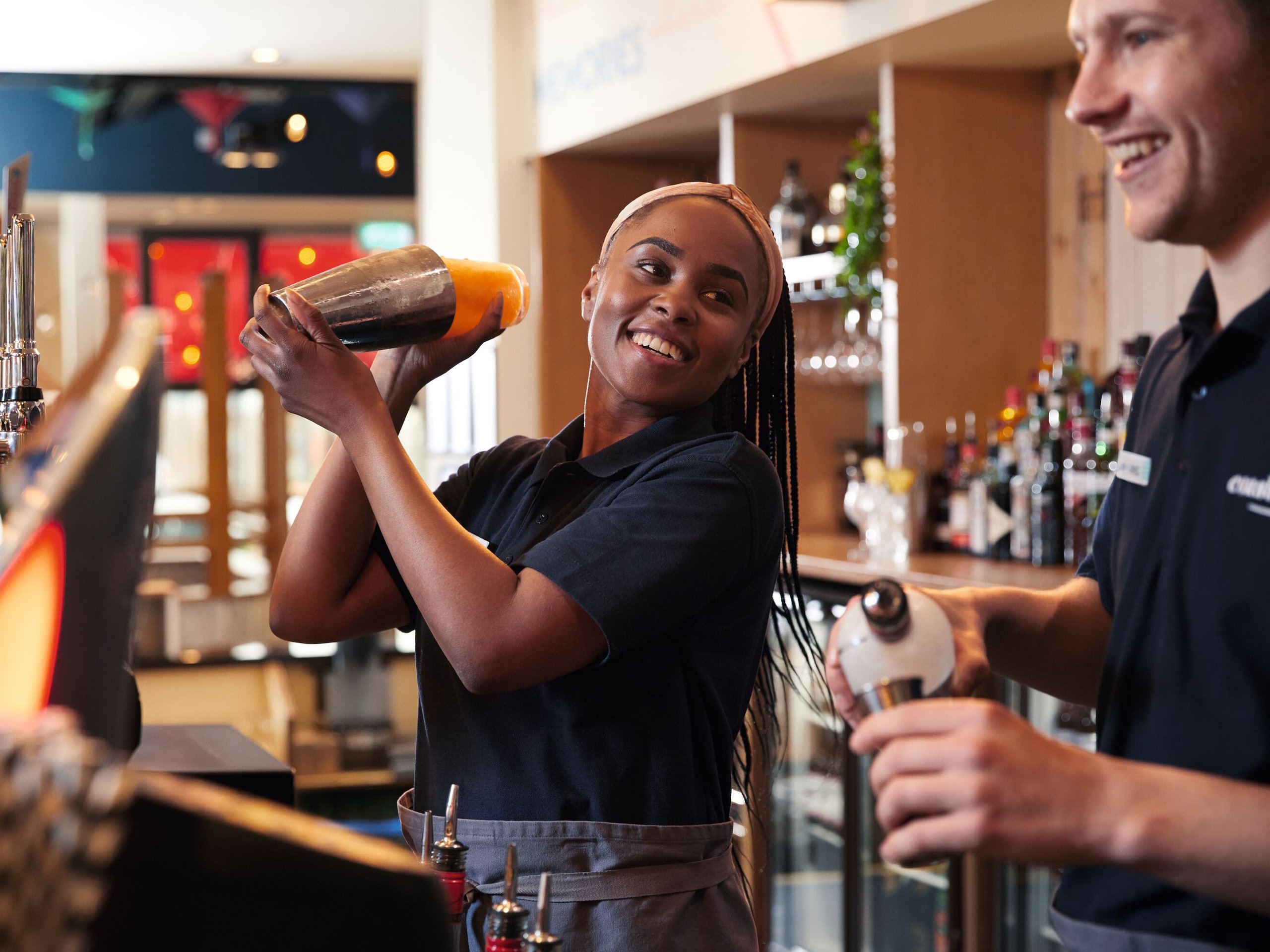 A Black woman with braids, wearing a navy polo shirt and apron, smiles as she shakes a cocktail shaker. A man in a similar uniform stands nearby.
