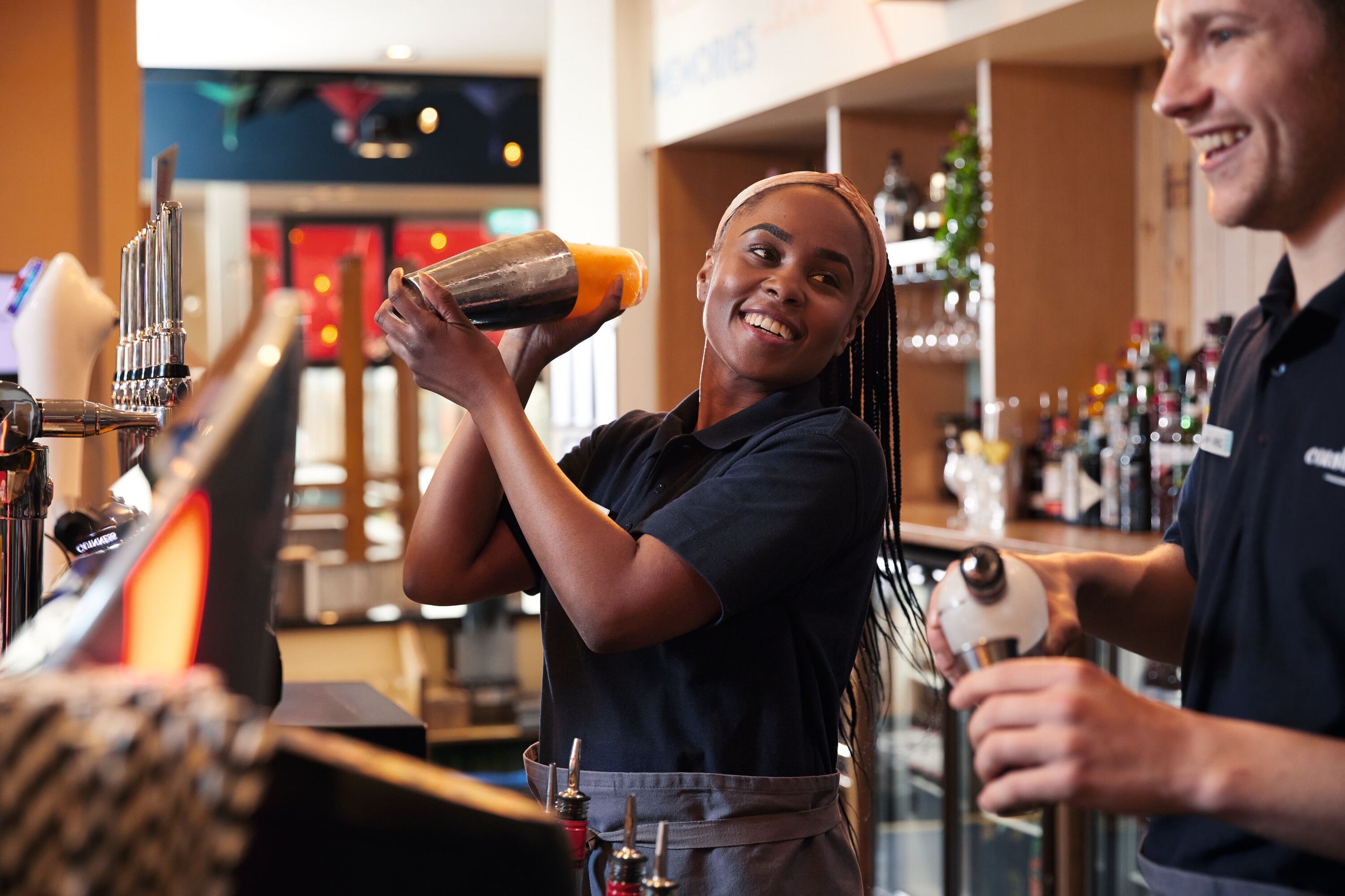 A Black woman with braids, wearing a navy polo shirt and apron, smiles as she shakes a cocktail shaker. A man in a similar uniform stands nearby.
