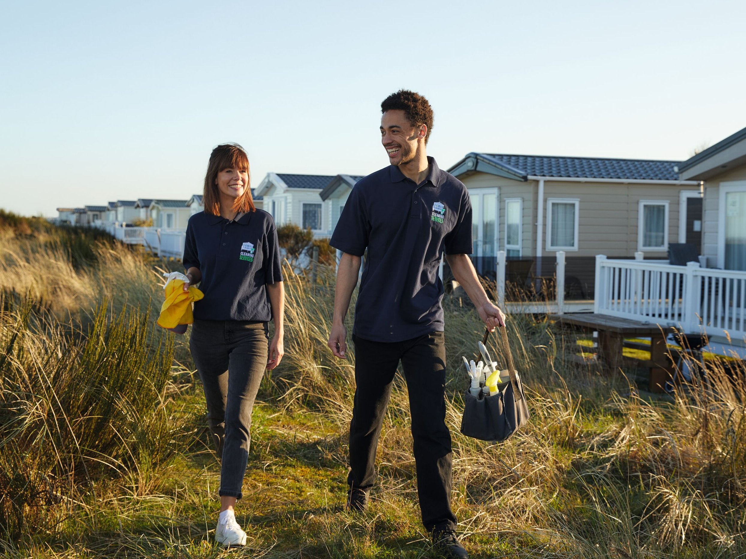 Two smiling cleaners in navy shirts walk past static caravans in a grassy area. One carries a yellow cloth and spray bottle, the other a cleaning caddy.