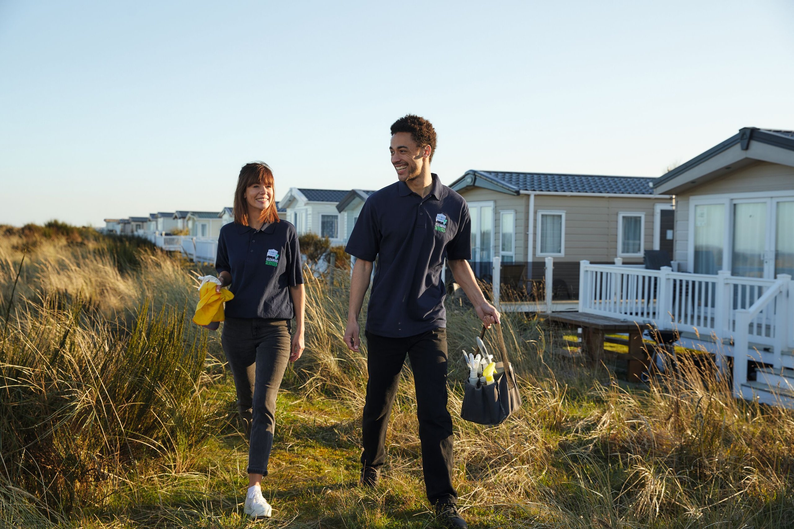 Two smiling cleaners in navy shirts walk past static caravans in a grassy area. One carries a yellow cloth and spray bottle, the other a cleaning caddy.