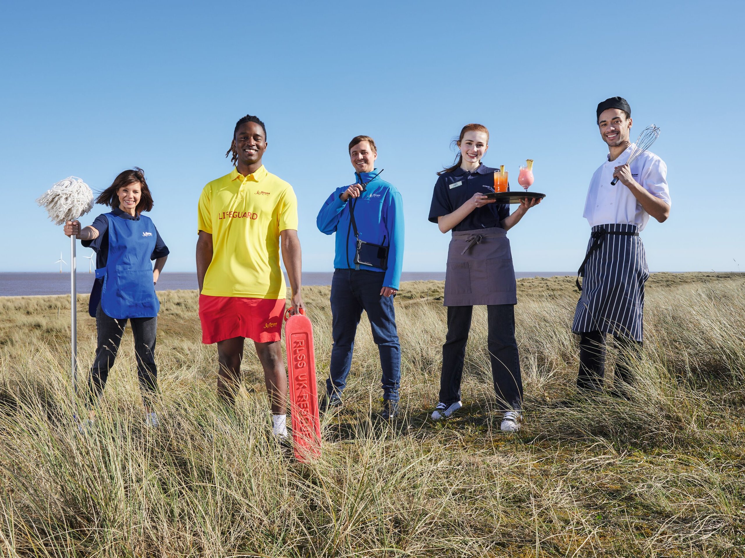 A team of five staff members, including a lifeguard, chef, waitress, and cleaner, stand on a grassy coastline with the sea and wind turbines in the background.
