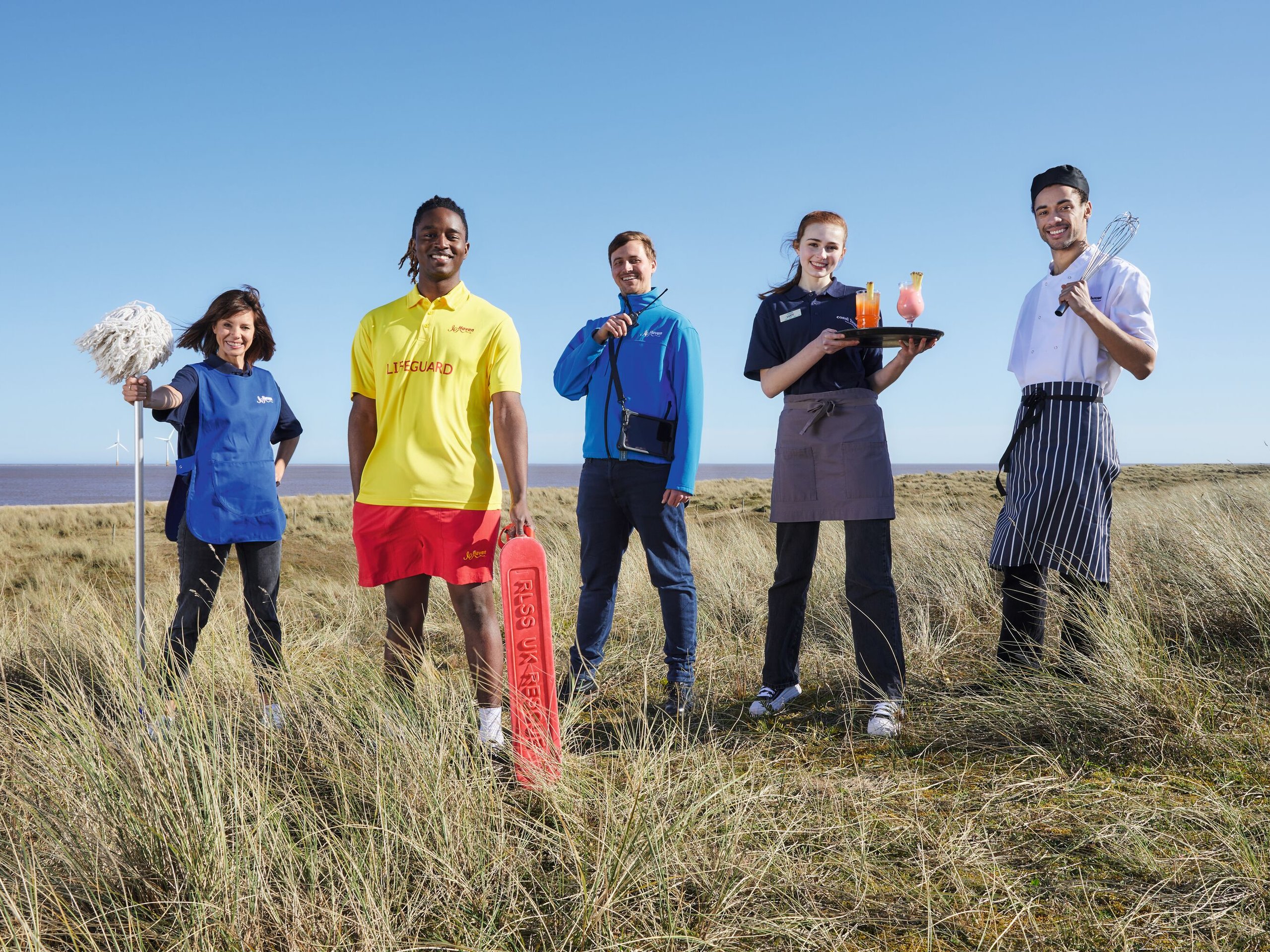 A team of five staff members, including a lifeguard, chef, waitress, and cleaner, stand on a grassy coastline with the sea and wind turbines in the background.