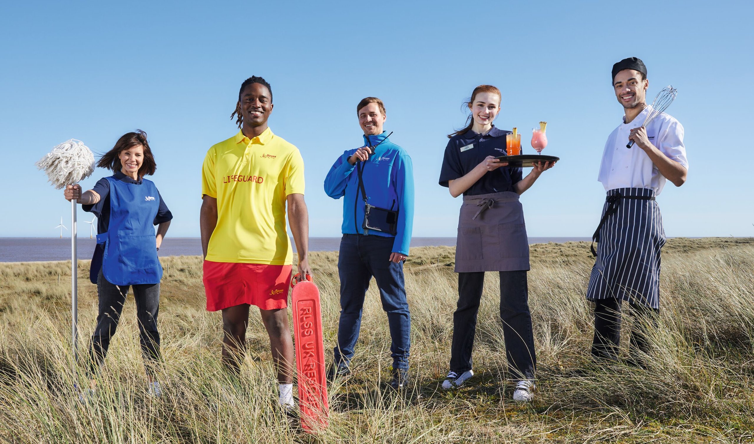 A team of five staff members, including a lifeguard, chef, waitress, and cleaner, stand on a grassy coastline with the sea and wind turbines in the background.