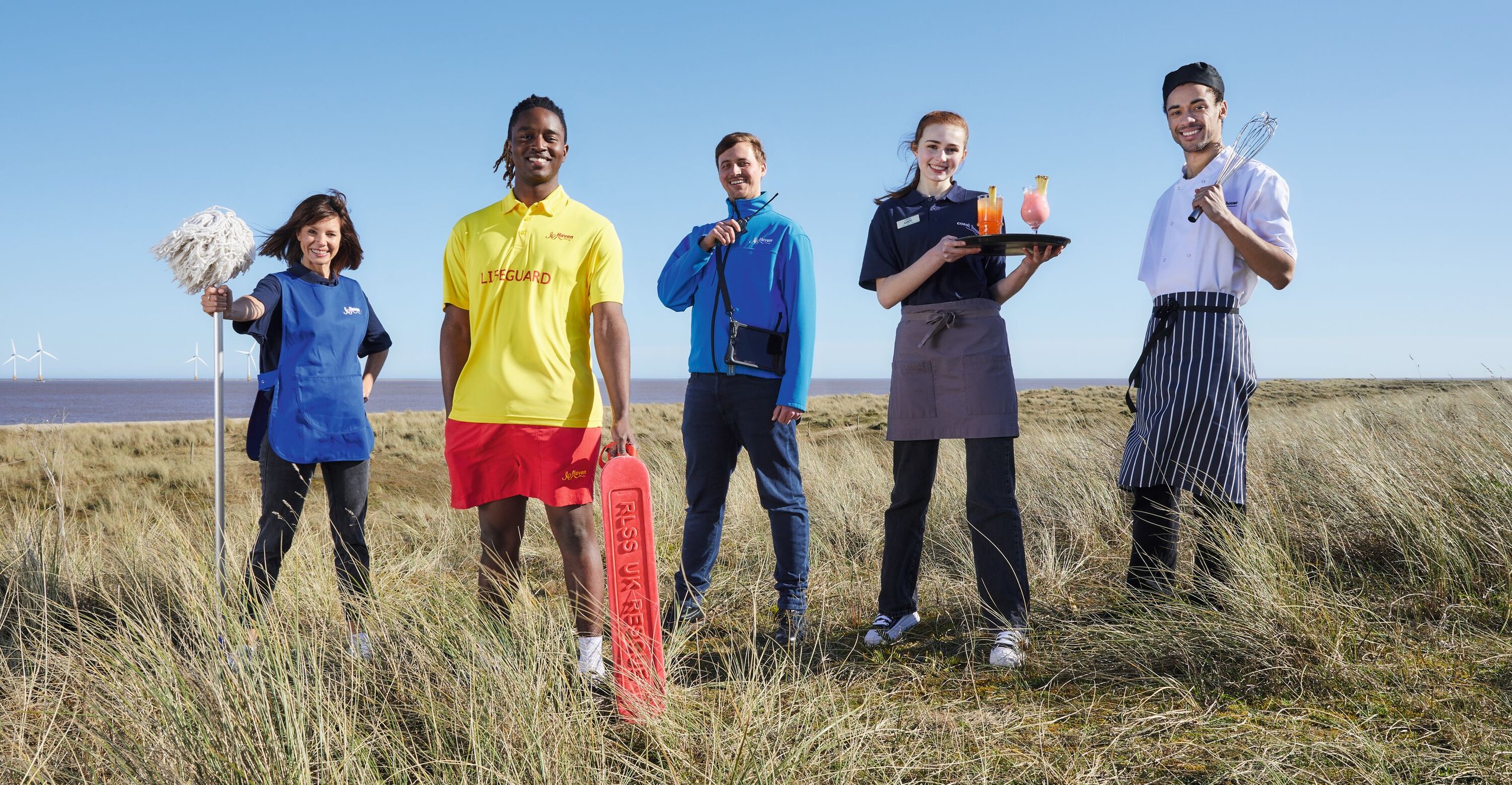 A team of five staff members, including a lifeguard, chef, waitress, and cleaner, stand on a grassy coastline with the sea and wind turbines in the background.