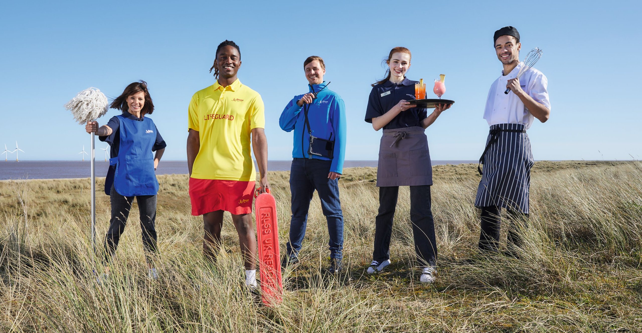 A team of five staff members, including a lifeguard, chef, waitress, and cleaner, stand on a grassy coastline with the sea and wind turbines in the background.