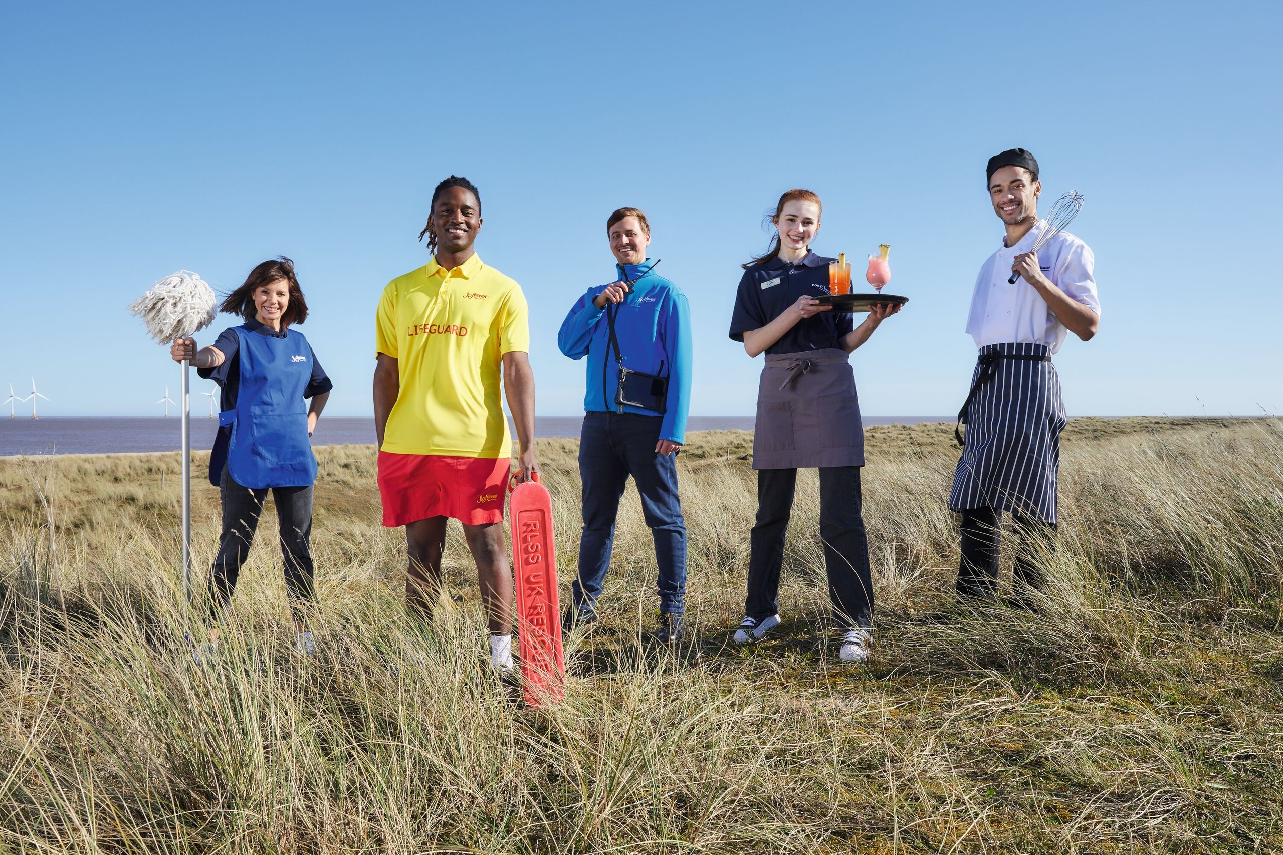 A team of five staff members, including a lifeguard, chef, waitress, and cleaner, stand on a grassy coastline with the sea and wind turbines in the background.