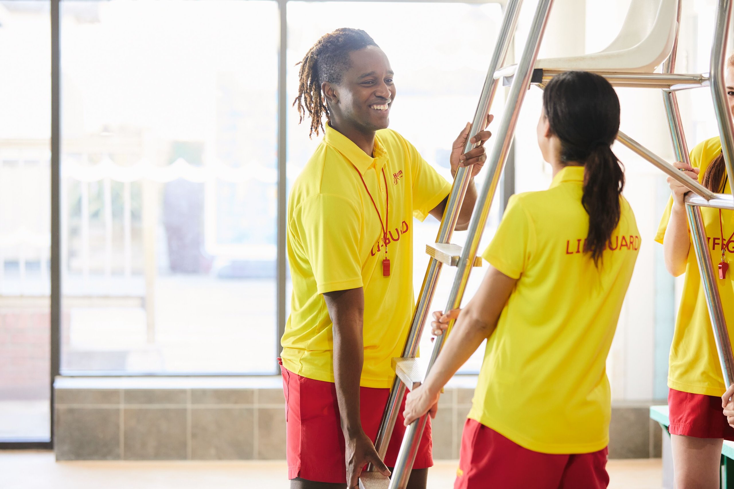 Three lifeguards in yellow shirts and red shorts stand near a lifeguard chair. One man smiles while holding the ladder.