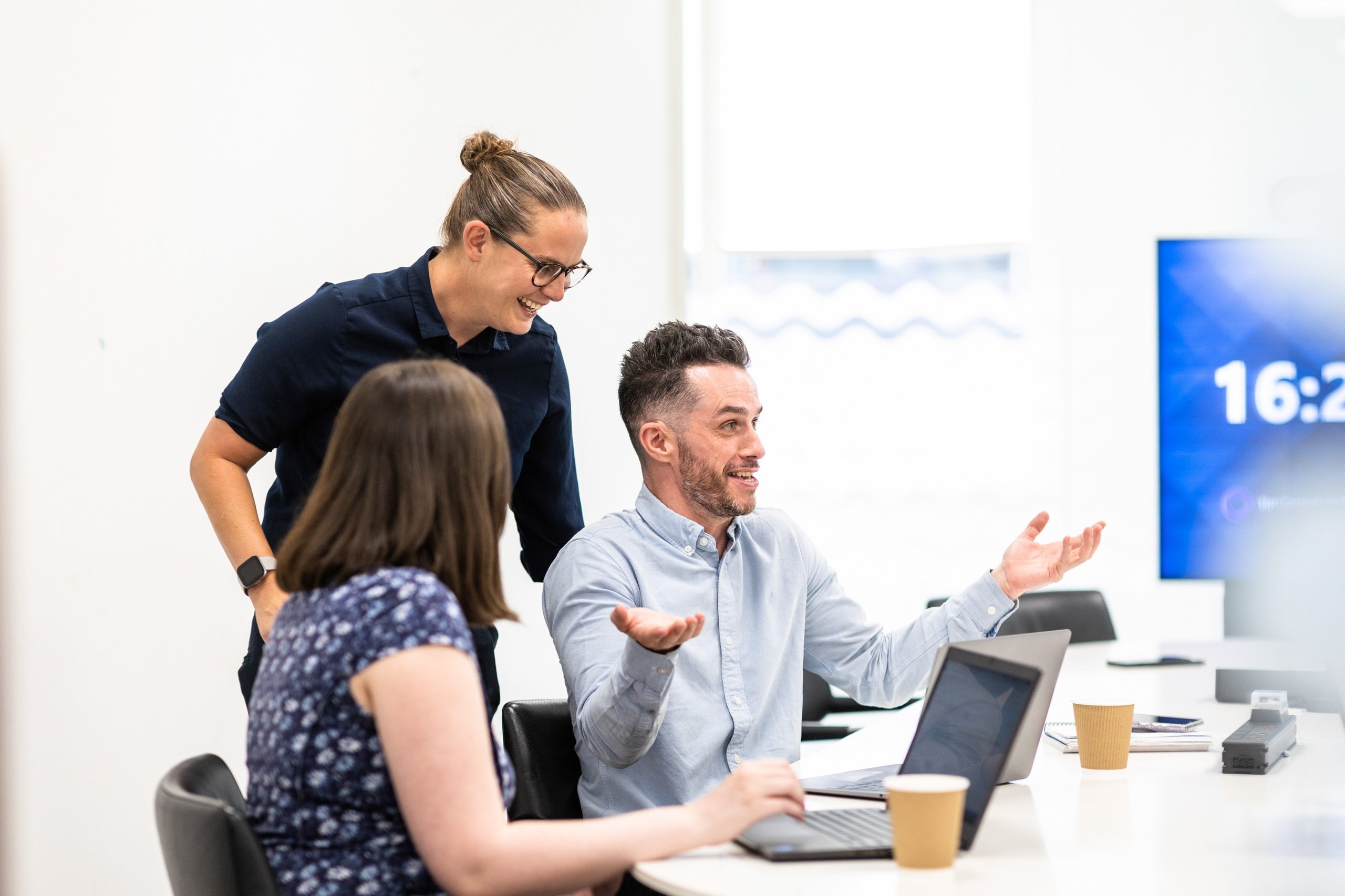 Three colleagues in a bright office, engaged in a discussion around a table with laptops and coffee cups.