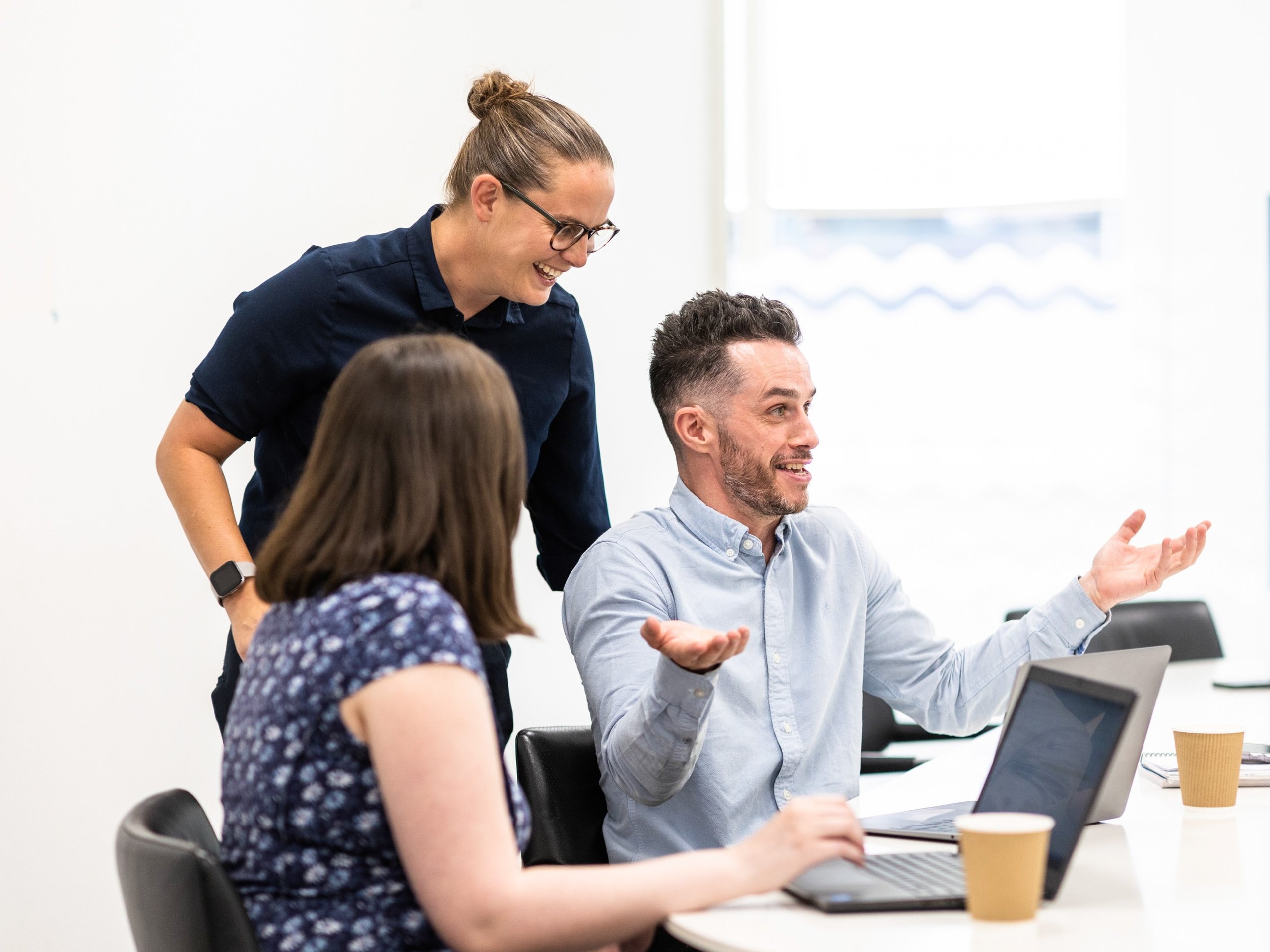 Three colleagues in a bright office, engaged in a discussion around a table with laptops and coffee cups.