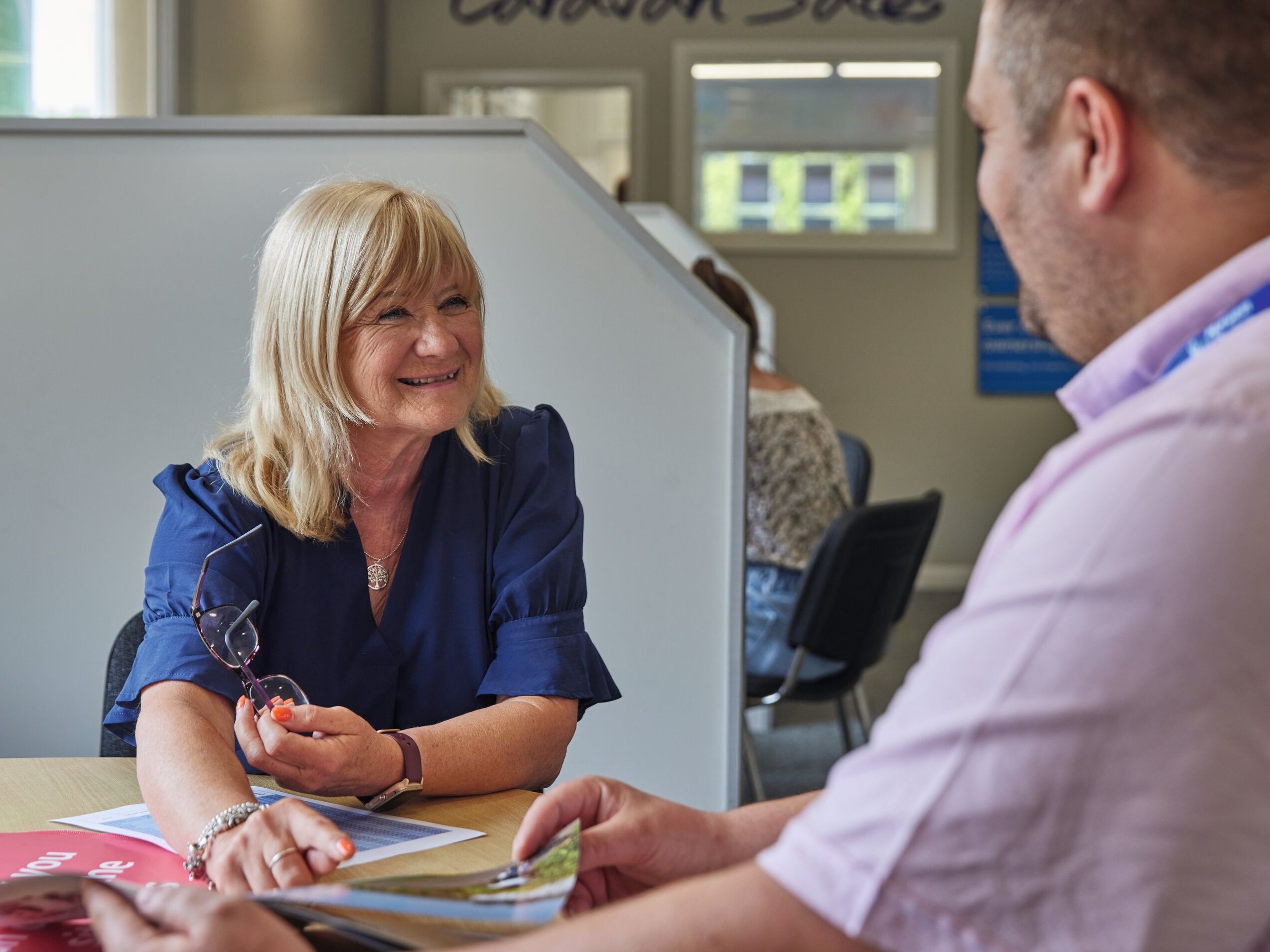 A woman in a blue shirt smiles as she talks to a man in a pink shirt, both sitting at a desk with papers.