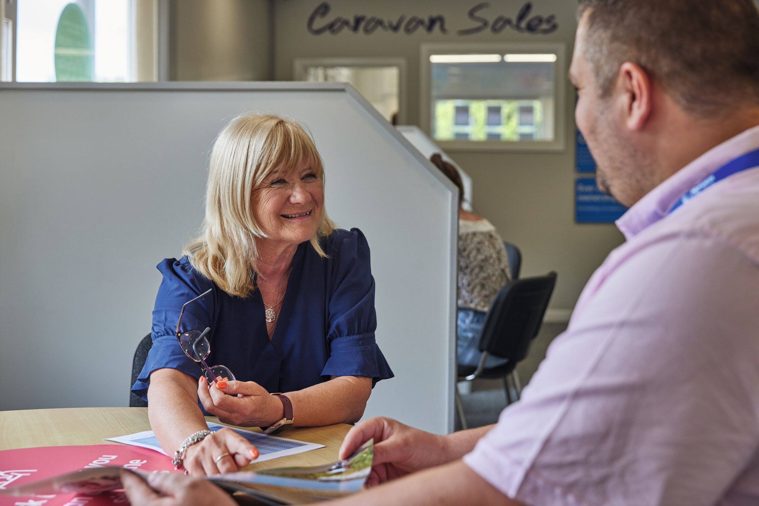A woman in a blue shirt smiles as she talks to a man in a pink shirt, both sitting at a desk with papers.