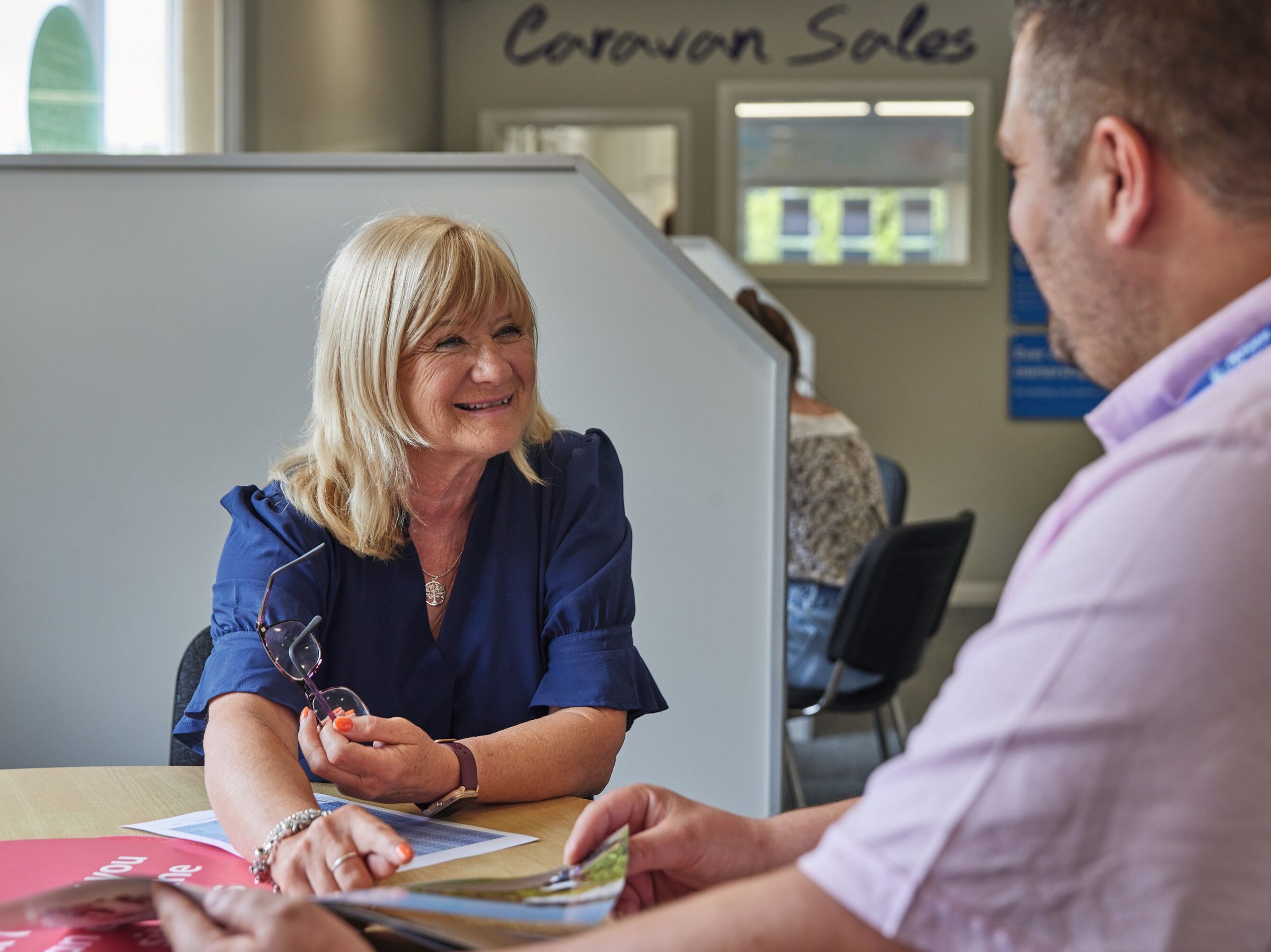 A woman in a blue shirt smiles as she talks to a man in a pink shirt, both sitting at a desk with papers.