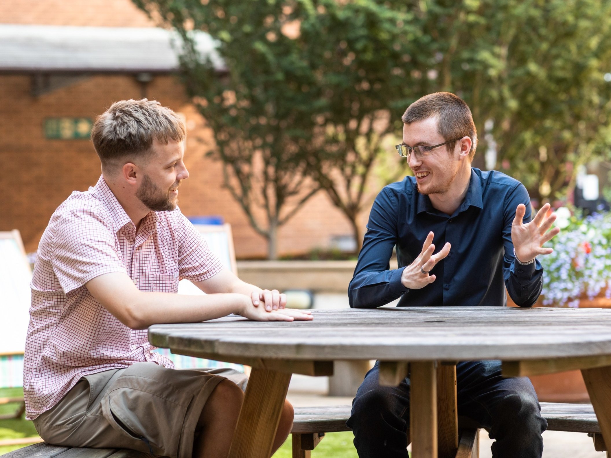 Two men in casual attire are sitting at a picnic table, engaged in conversation in an outdoor setting with greenery and a brick building in the background.