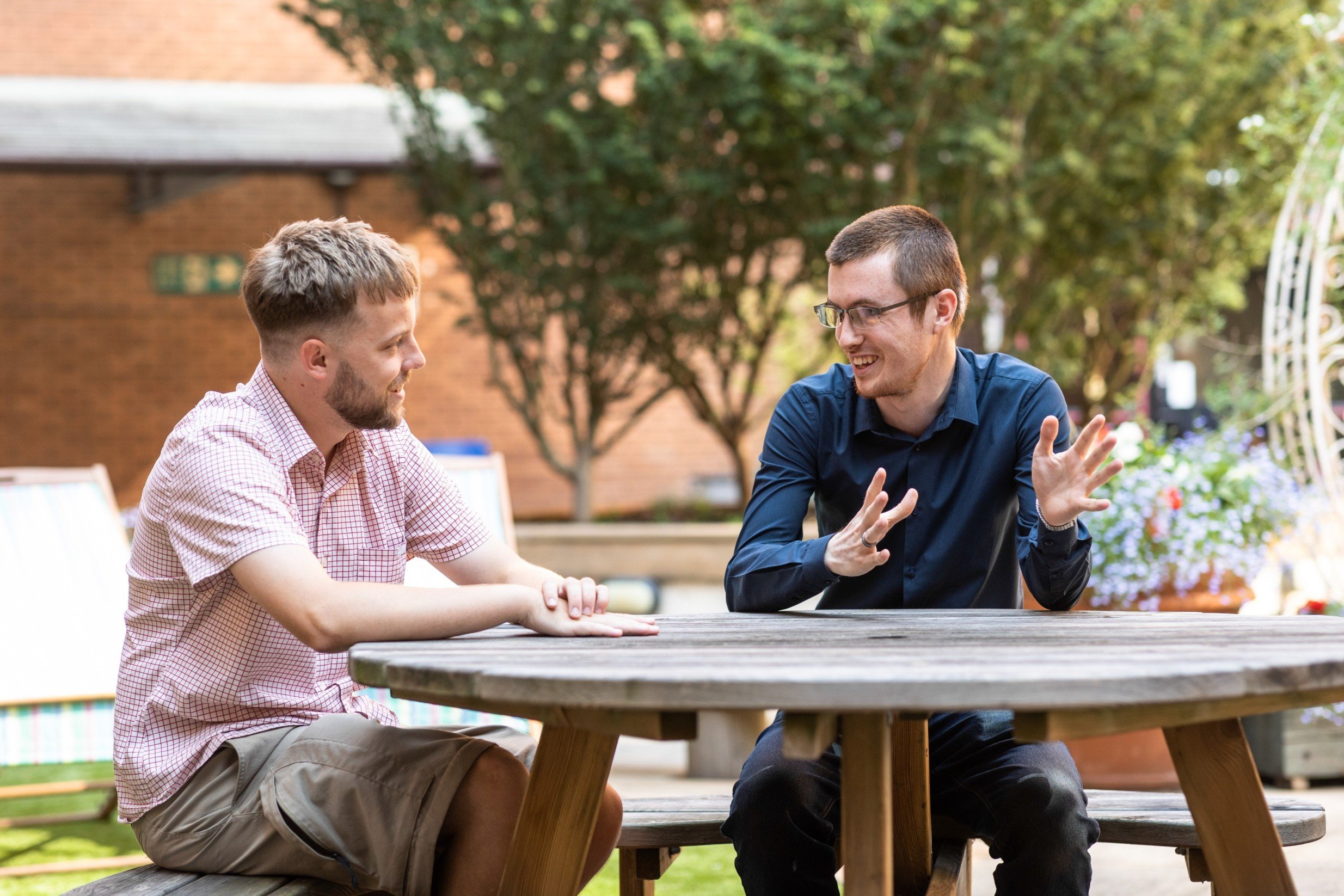 Two men in casual attire are sitting at a picnic table, engaged in conversation in an outdoor setting with greenery and a brick building in the background.