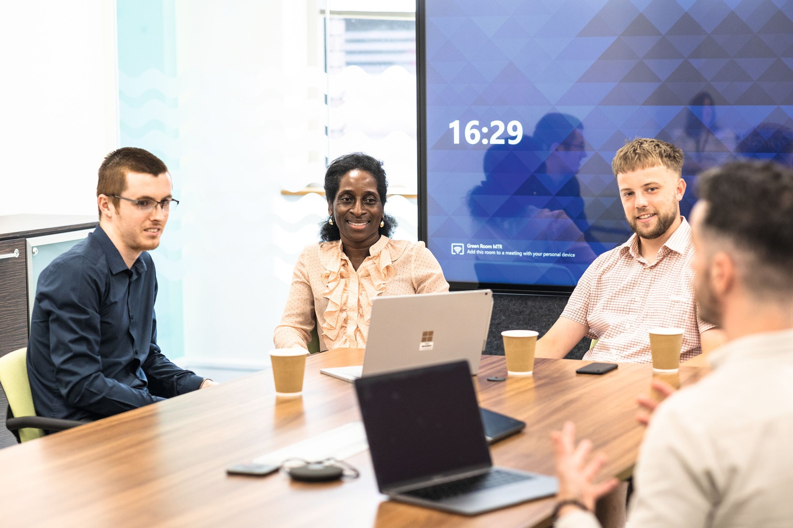 Four professionals in a bright office participate in a meeting around a wooden table. A large screen displays the time and a message about the meeting room.