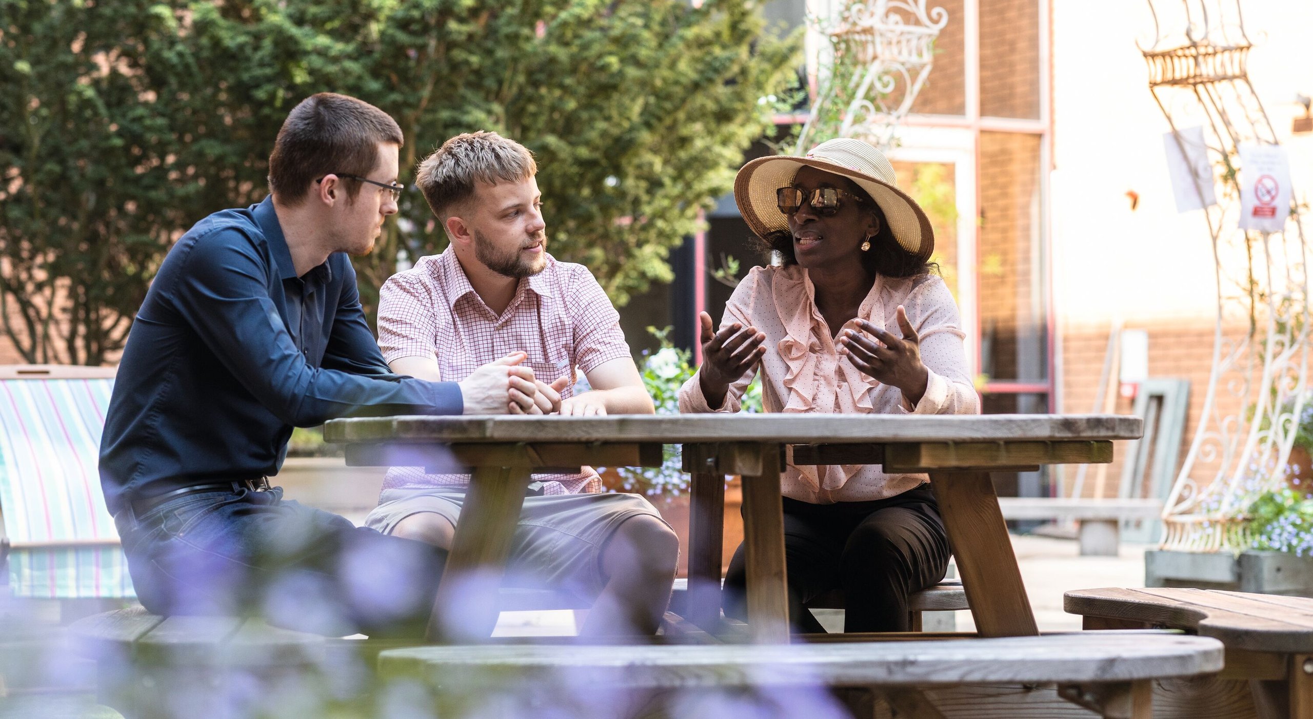 Three colleagues, two men and a woman in a hat and sunglasses, sit at a picnic table outdoors, engaged in conversation.
